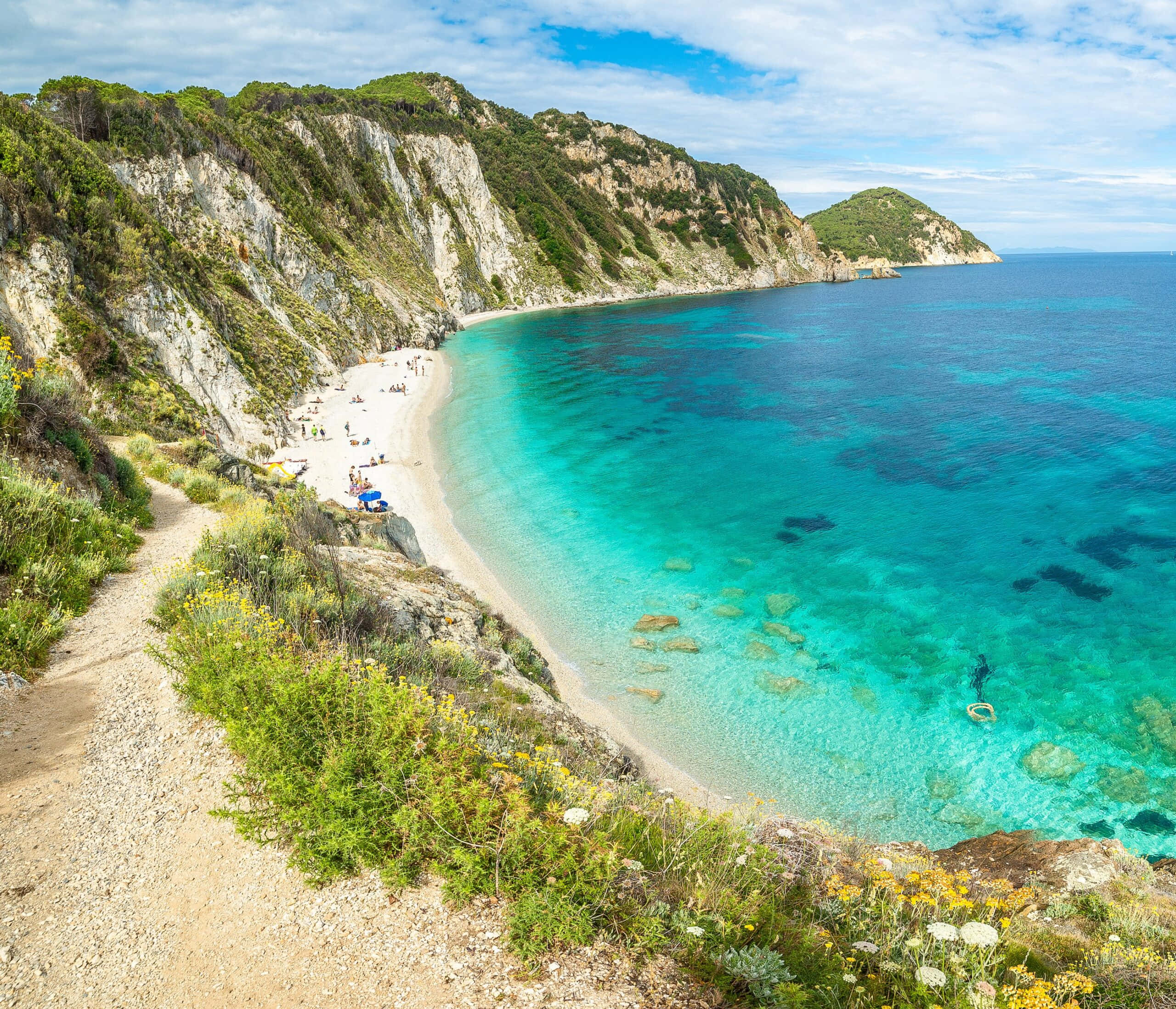 Italian Beach 桌布 壁紙 — 一個令人驚嘆的海岸小海灣，碧綠的海水擁抱著沙灘，沙灘兩旁是鬱鬱蔥蔥的綠色懸崖，上方是明亮的藍天。