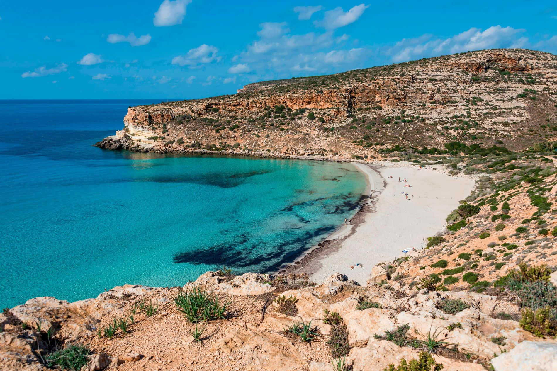 Italian Beach 桌布 壁紙 — 一個令人驚嘆的海灣，碧綠的海水環繞著白色沙灘，沙灘坐落在崎嶇的懸崖之間，上方是明亮的藍天。