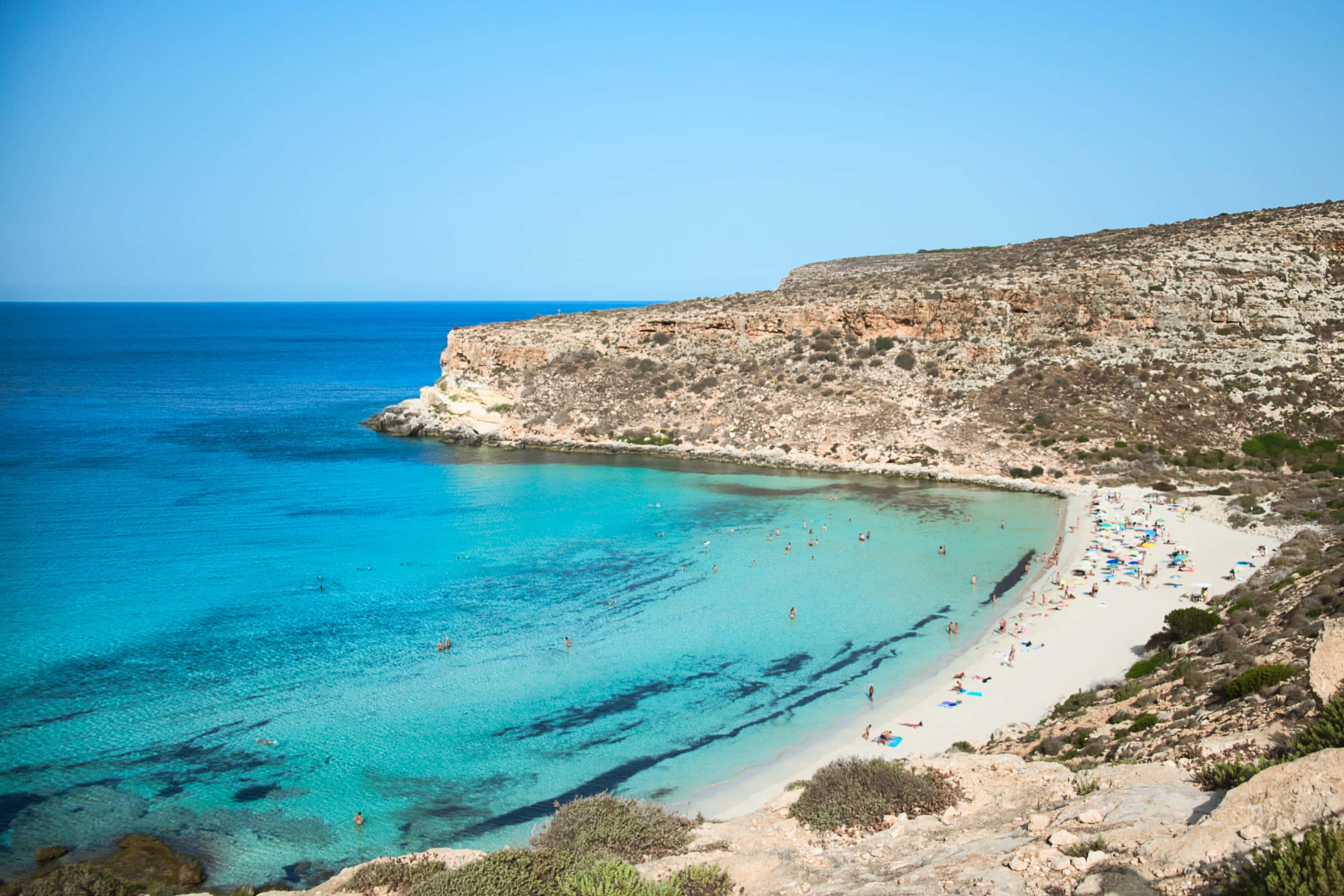 Mediterranean Beach 桌布 壁紙 — 一個令人驚嘆的碧綠海灣，擁有白色沙灘，隱藏在崎嶇的懸崖之間，天空清澈湛藍。