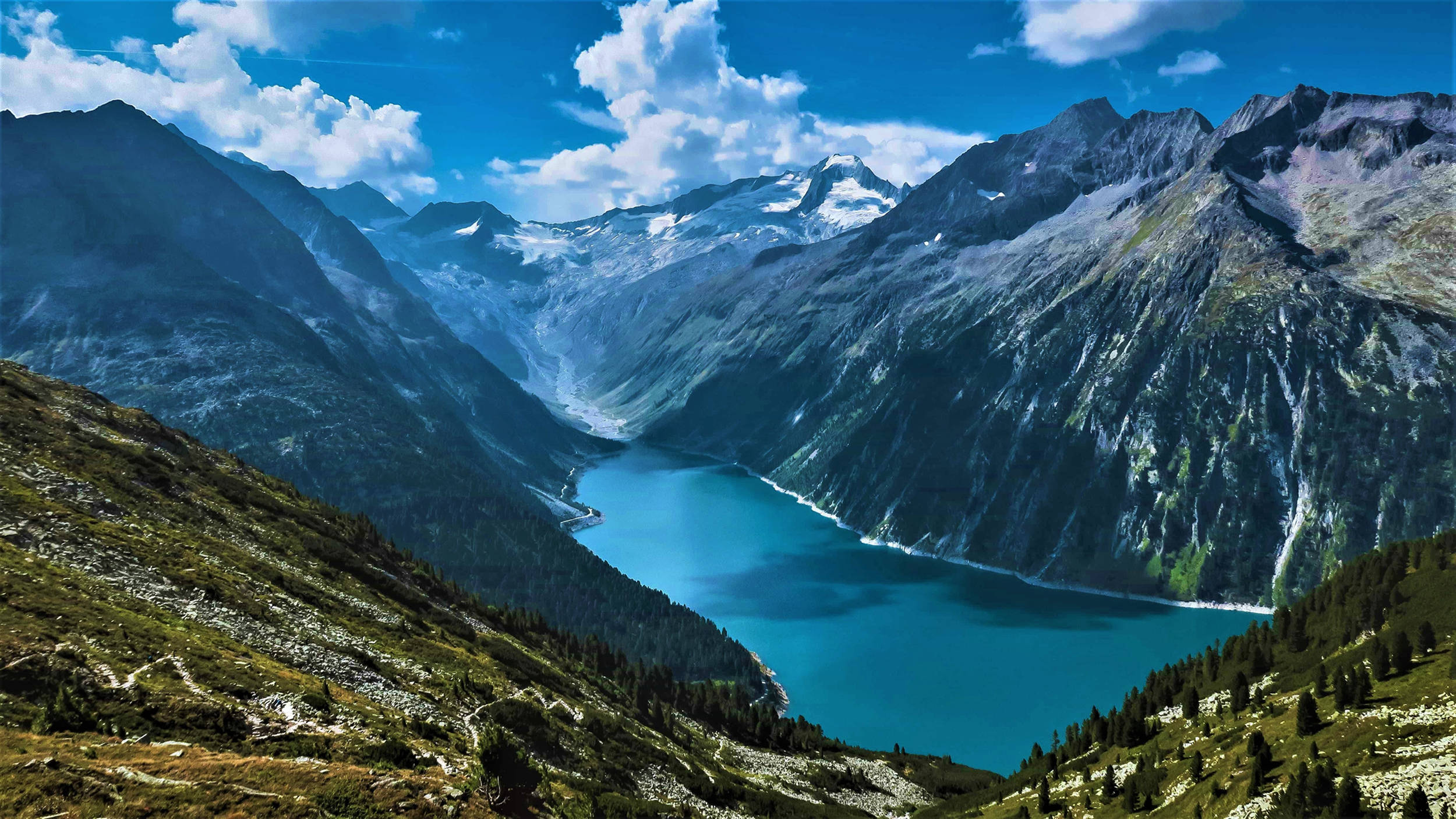 Norway 桌布 壁紙 — 一個令人驚嘆的高山湖泊，坐落在高聳的雪山之間，天空湛藍，白雲朵朵。