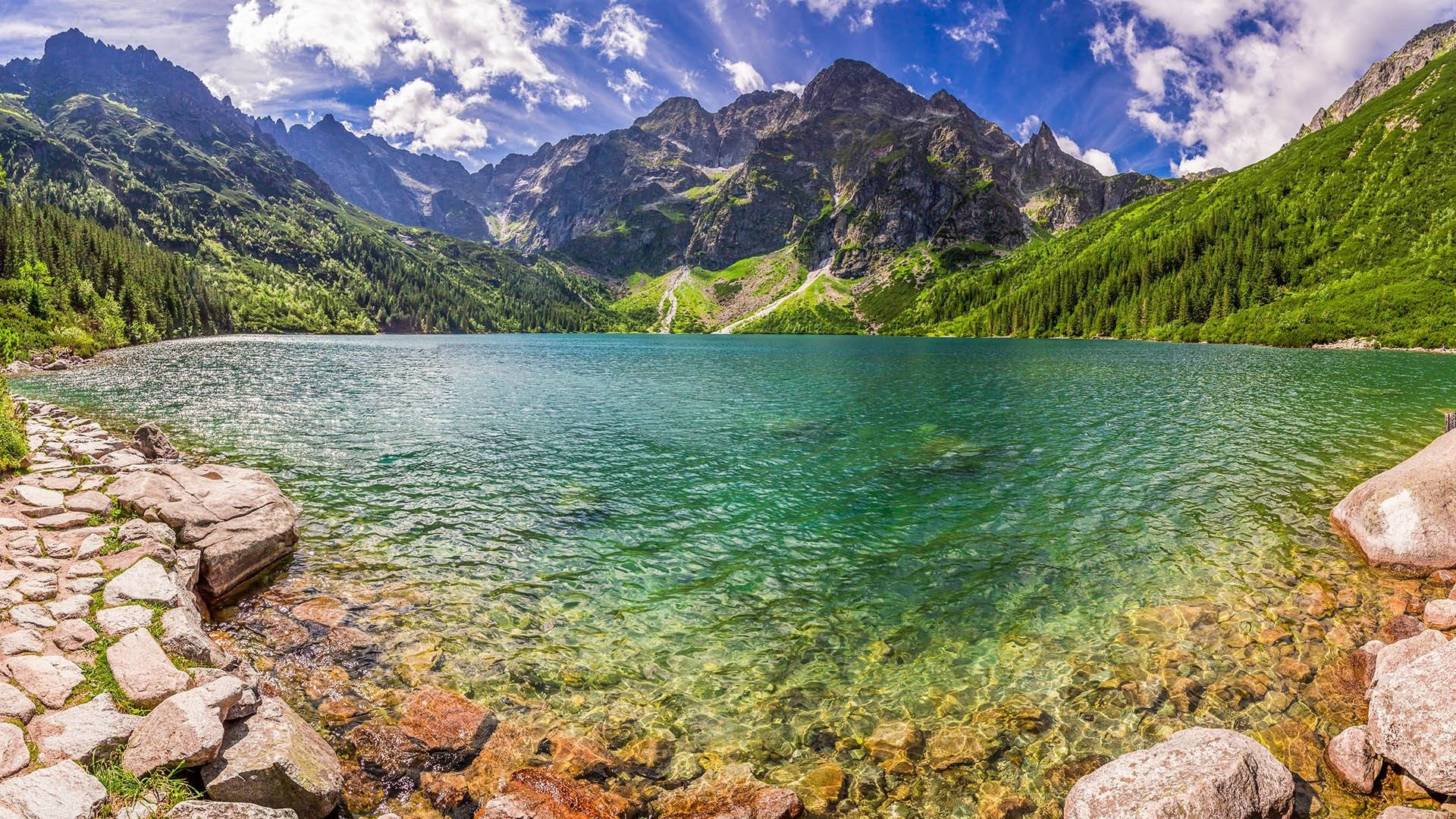 在明亮的藍天下，一個清澈碧綠的湖泊坐落在鬱鬱蔥蔥的綠色山脈之間。 — 波蘭 GIF 壁紙