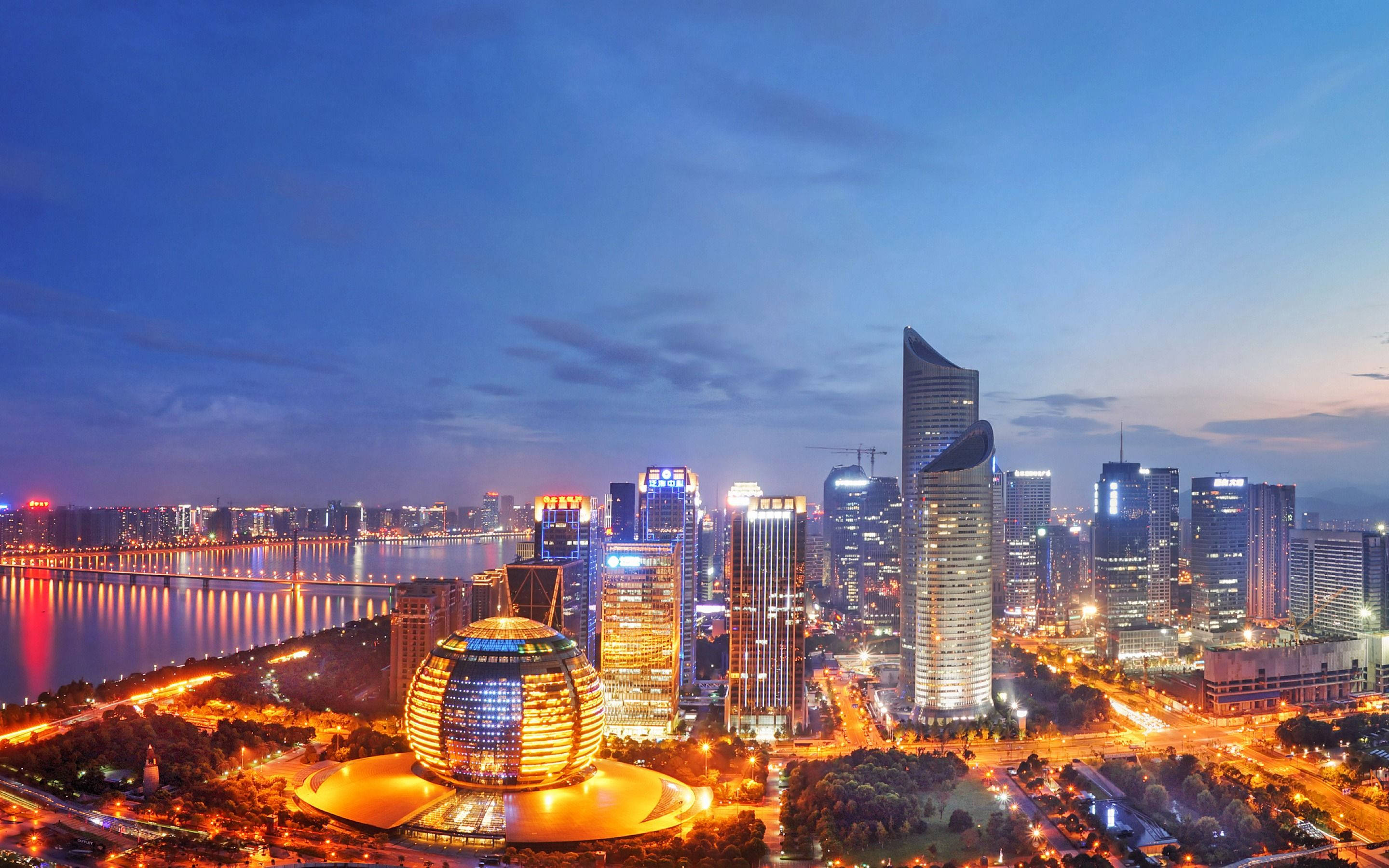 Hangzhou 桌布 壁紙 — 一個令人驚嘆的黃昏城市景觀，閃爍的摩天大樓和一條在深藍色天空下反射溫暖燈光的河流。