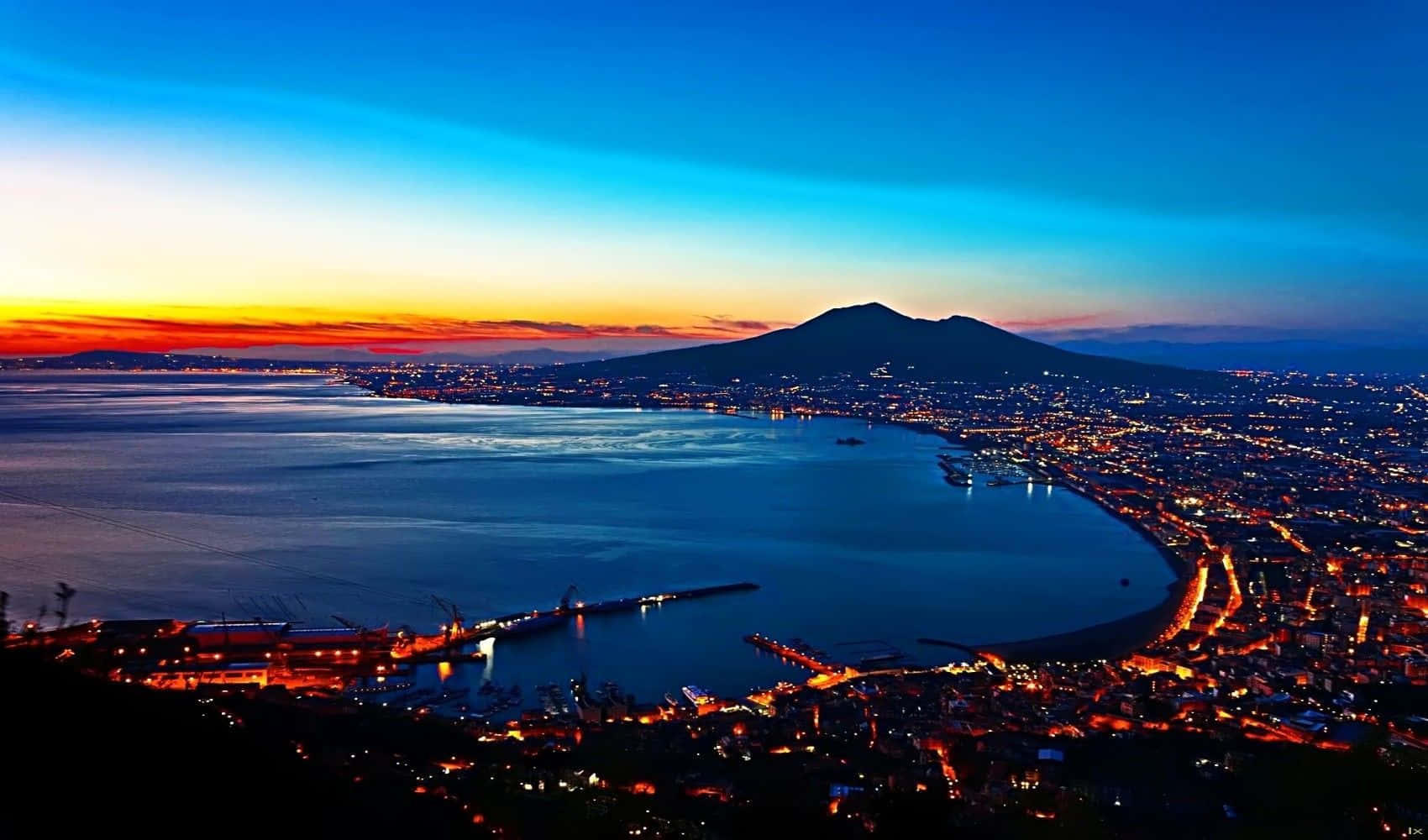 Volcano 桌布 壁紙 — 一個令人驚嘆的黃昏景色，海岸城市環繞海灣而建，遠處有一座顯著的火山。