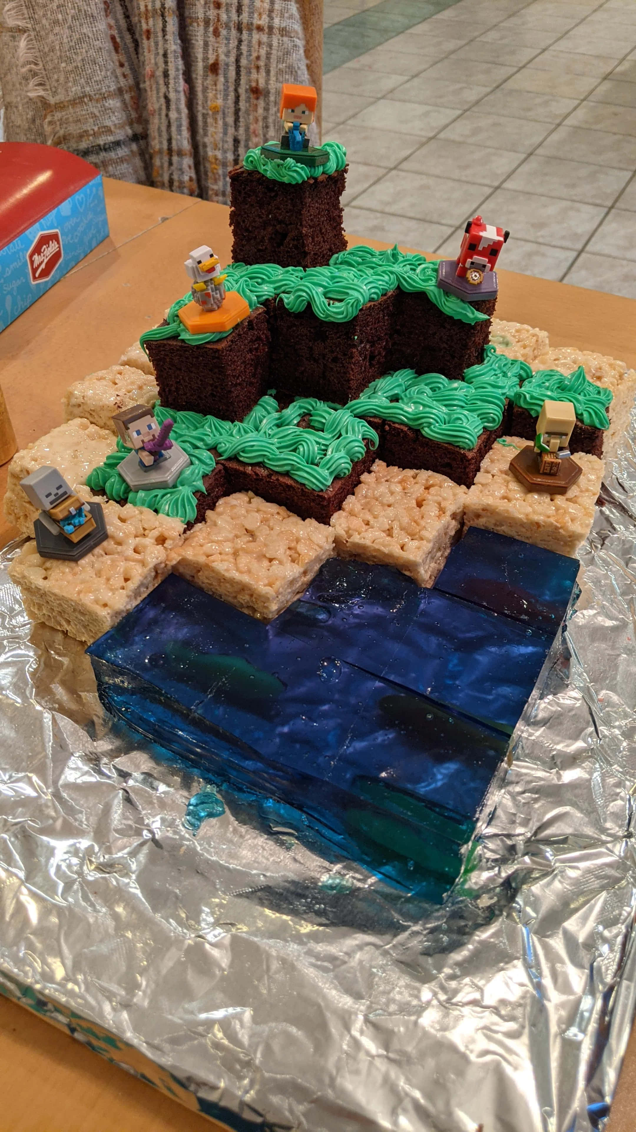 Minecraft Cakes 桌布 壁紙 — 一個以Minecraft為主題的蛋糕，由Rice Krispies零食和巧克力塊製成，帶有綠色糖霜河流和小角色公仔。