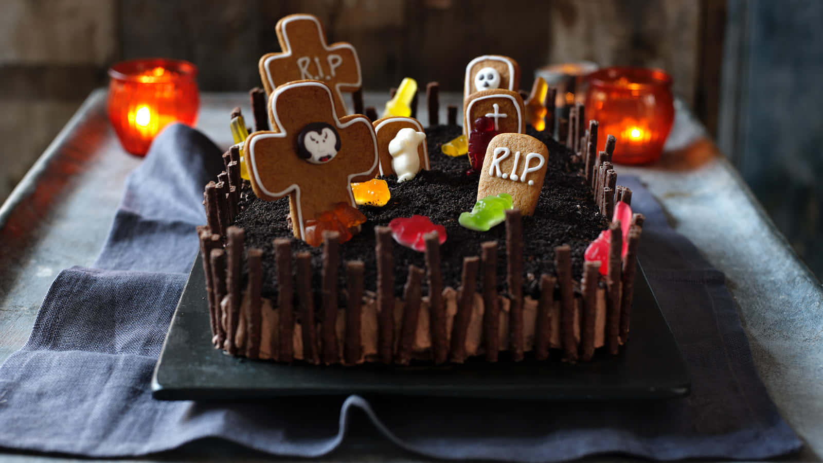 Halloween Cake 桌布 壁紙 — 一個以墓地為造型的詭異萬聖節蛋糕，配有薑餅墓碑、軟糖蟲和巧克力圍欄，在溫暖燭光下。