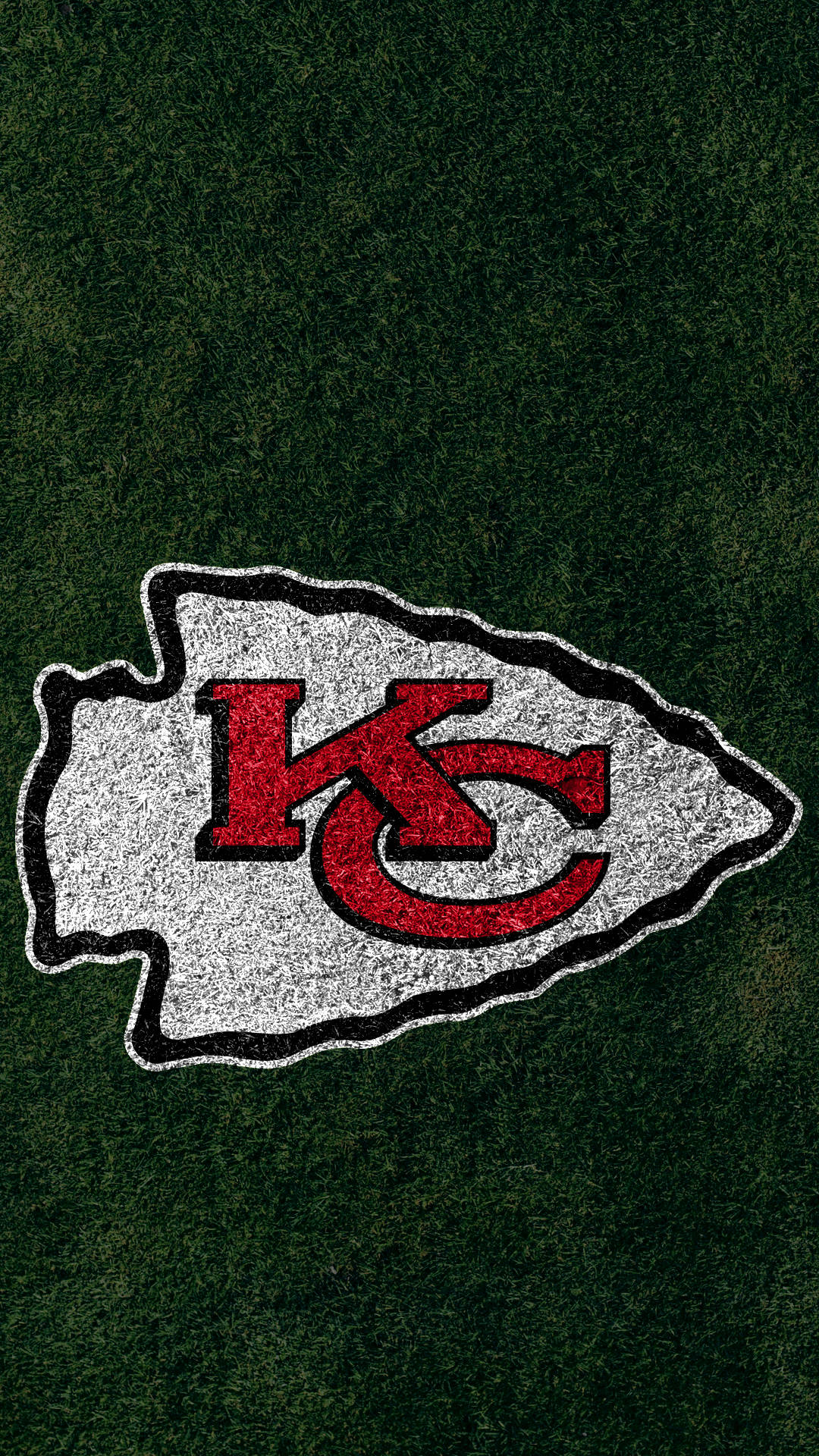 Kc Chiefs 桌布 壁紙 — 一個以紅色和銀色呈現的堪薩斯城酋長隊標誌，背景為帶有紋理的綠色草地。