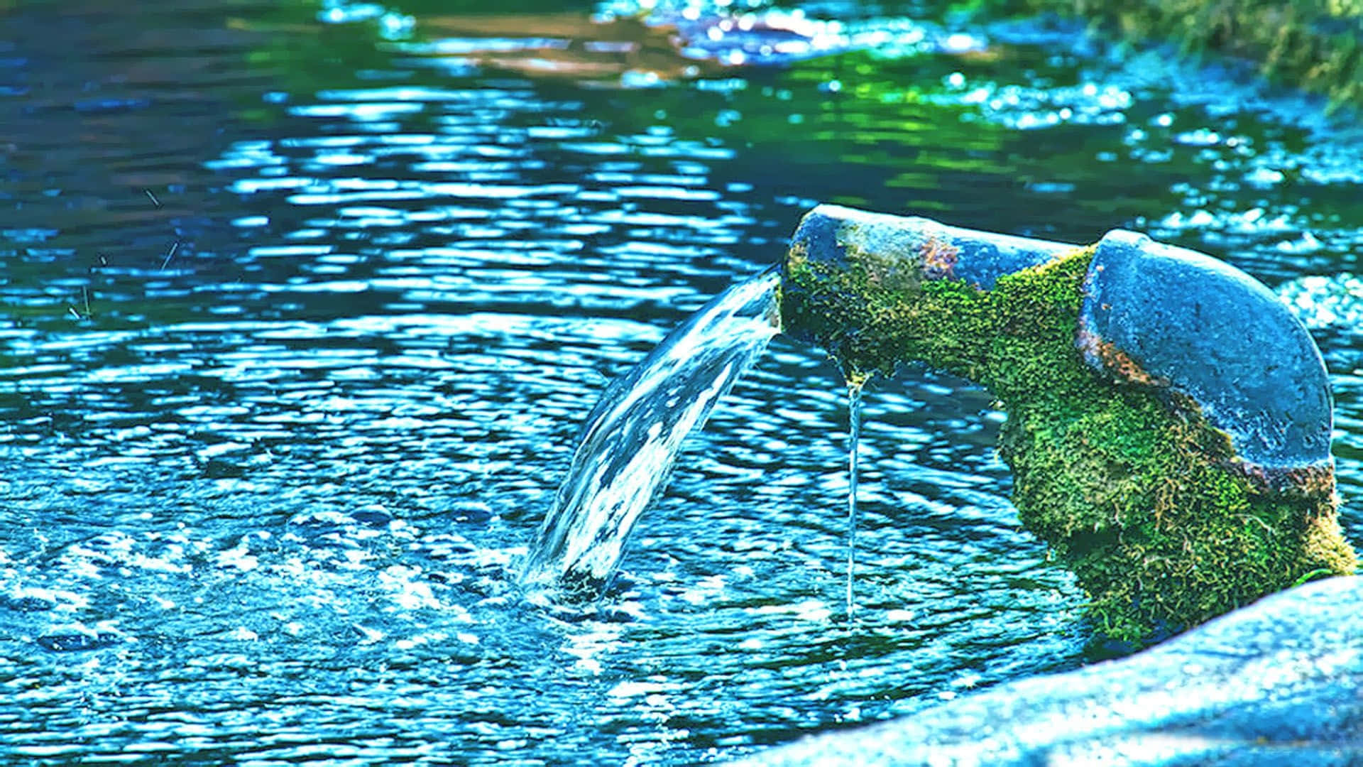 Spring Water 桌布 壁紙 — 一個佈滿青苔的金屬出水口將清澈的水倒入一個閃爍的藍色水池中，倒映著綠色的樹葉。