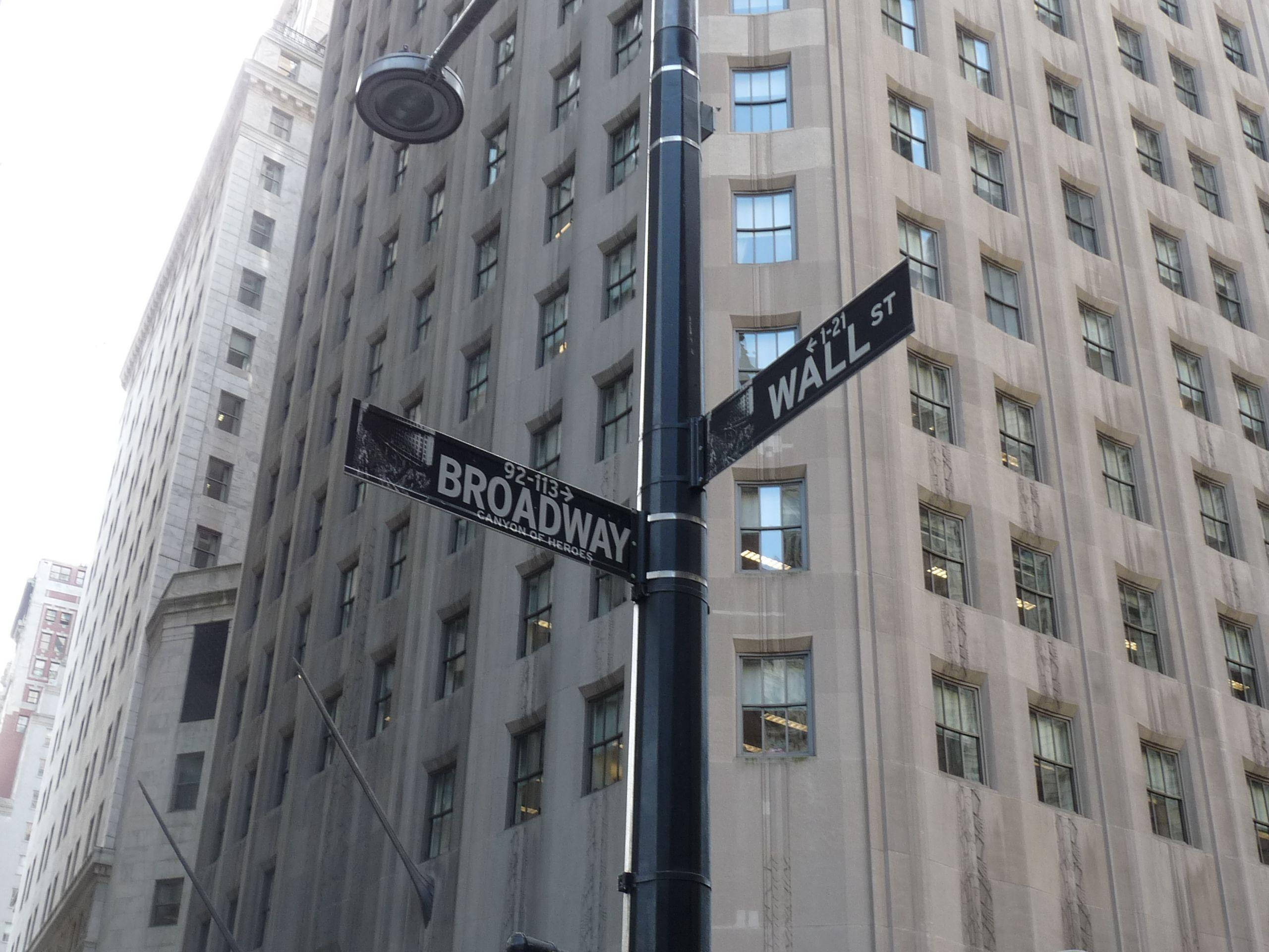 Broadway 桌布 壁紙 — 一個位於百老匯和華爾街交叉口的路牌，矗立在一座高聳的石造摩天大樓前，天空多雲。