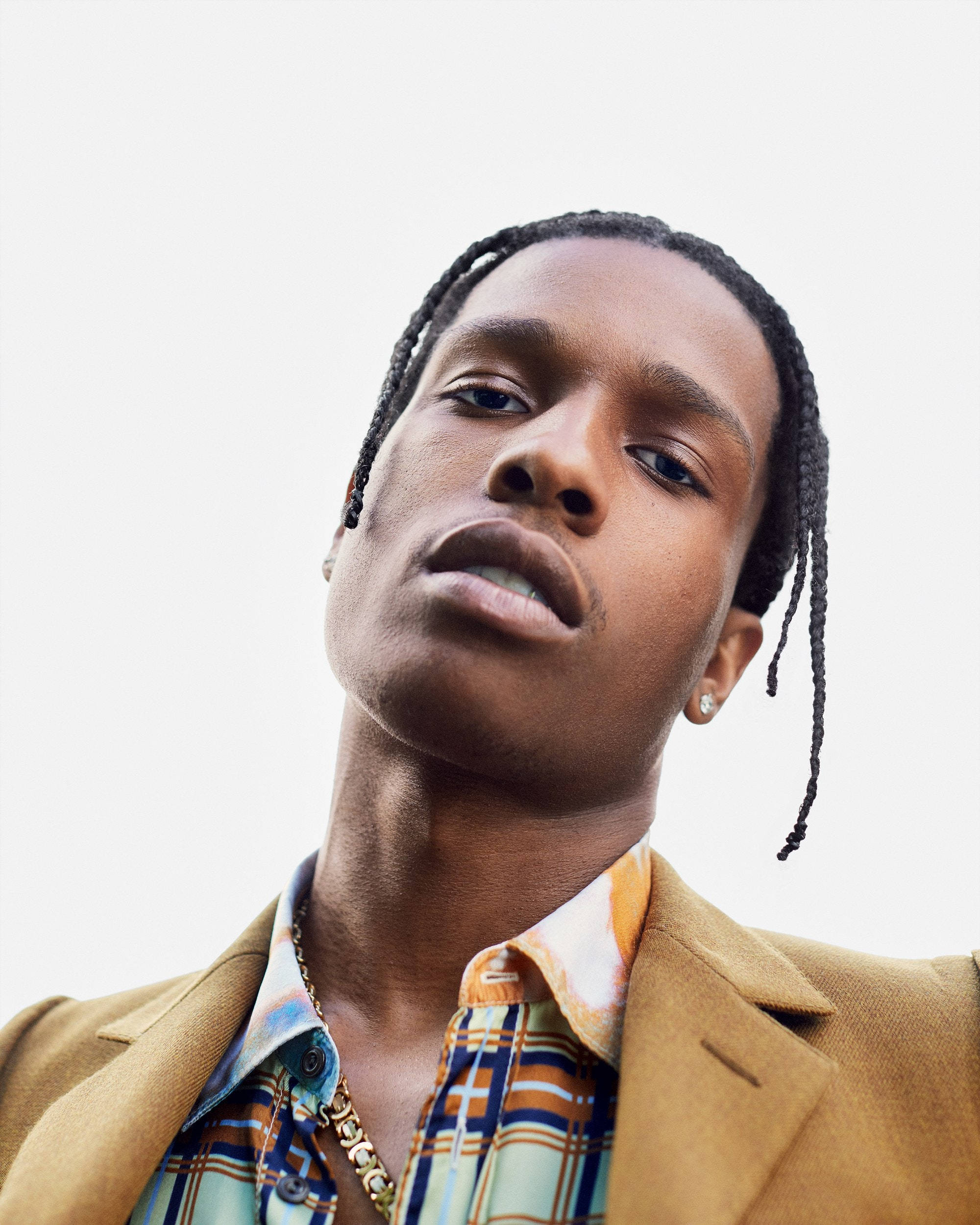 rocky 桌布 壁紙 — 一個低角度肖像，一位時尚男士編著辮子，身穿米色西裝外套和圖案襯衫，背景為純白色。