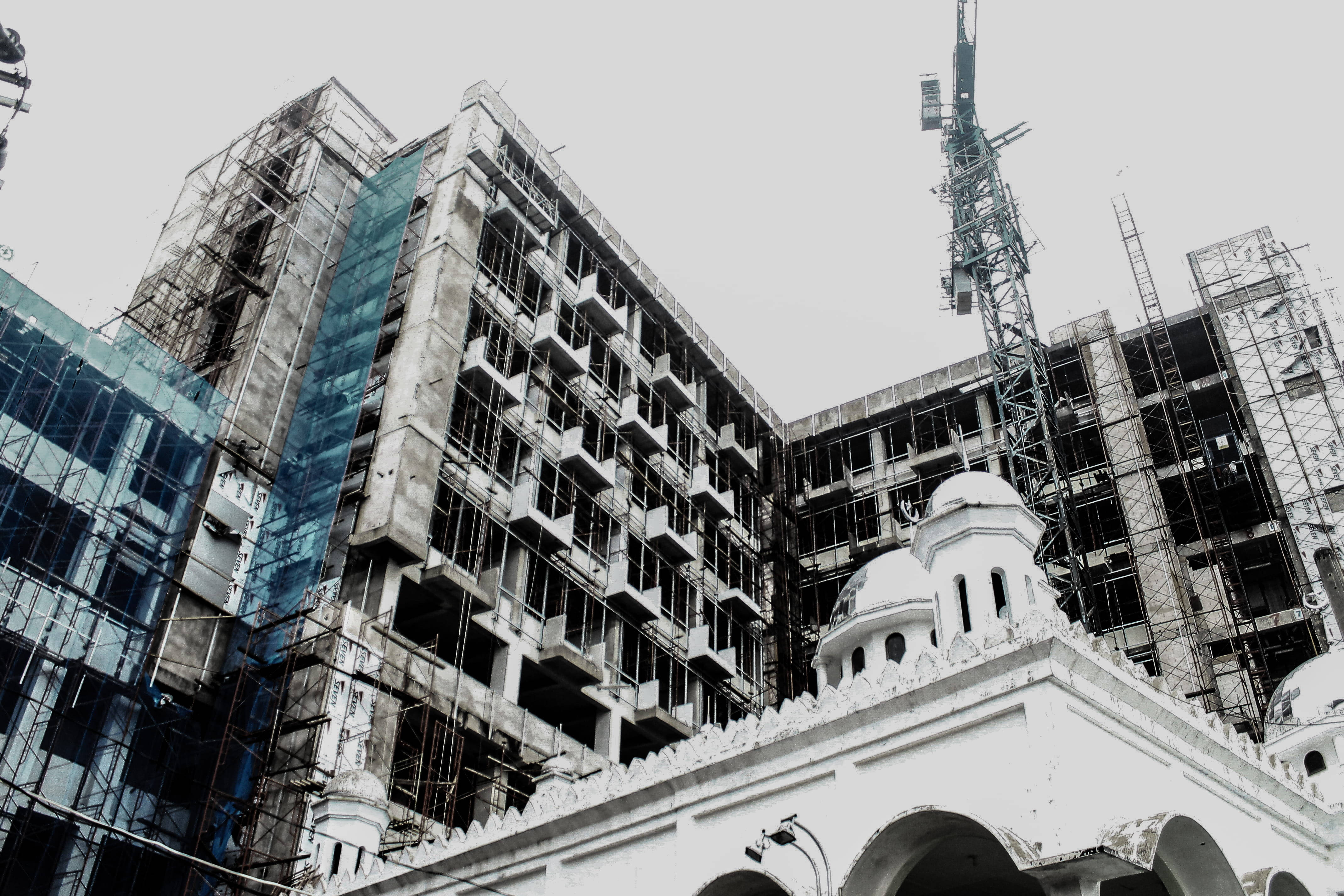 Architecture 桌布 壁紙 — 一個低角度視角，一座帶有圓頂的樸素白色清真寺，與一座被鷹架包裹著的未完工混凝土高樓在陰天天空下形成對比。