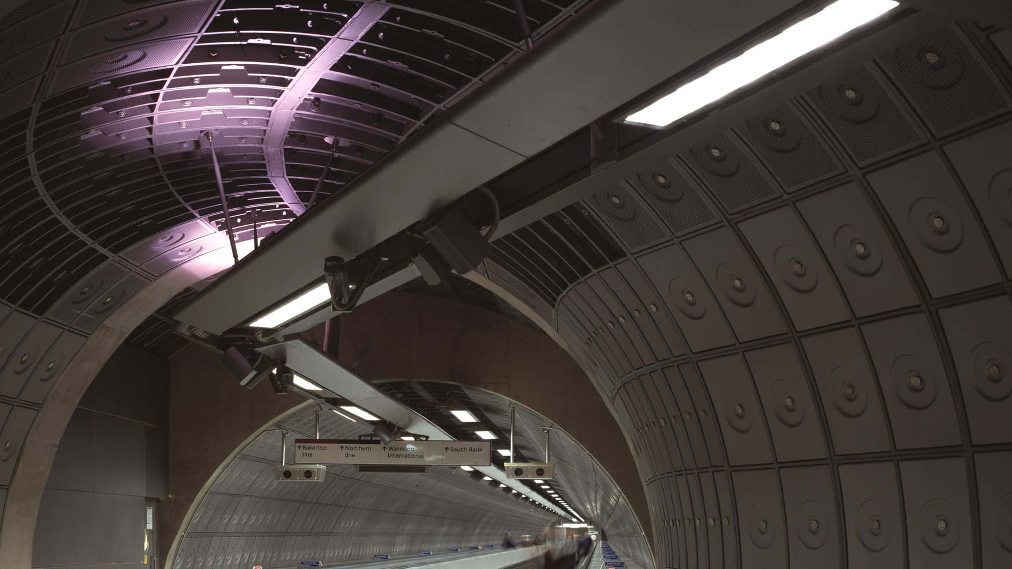train tunnel 桌布 壁紙 — 一個低角度視角，展示帶有金屬牆壁和紫色照明天花板面板的彎曲地鐵隧道。