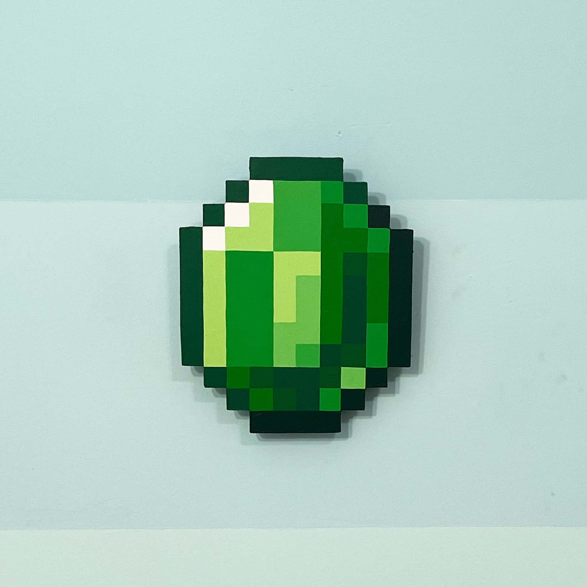 Minecraft Emeralds 桌布 壁紙 — 一個來自Minecraft的像素化綠色鑽石方塊安裝在淺藍色牆壁上，投射出柔和的陰影。