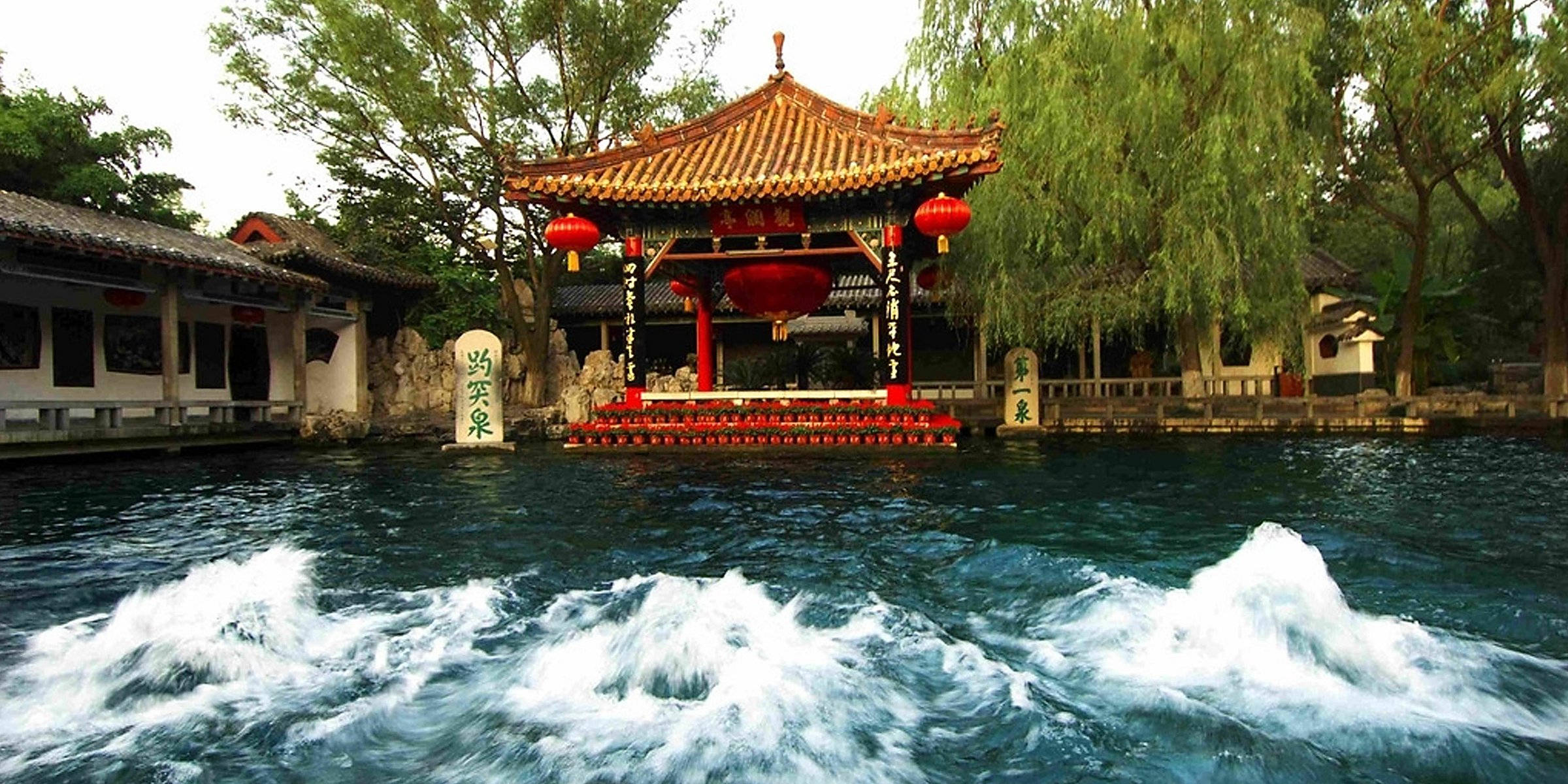 中國 桌布 壁紙 — 一個傳統中式亭台掛著紅燈籠，矗立在湍急的泉水池上，四周環繞著茂密的綠樹。