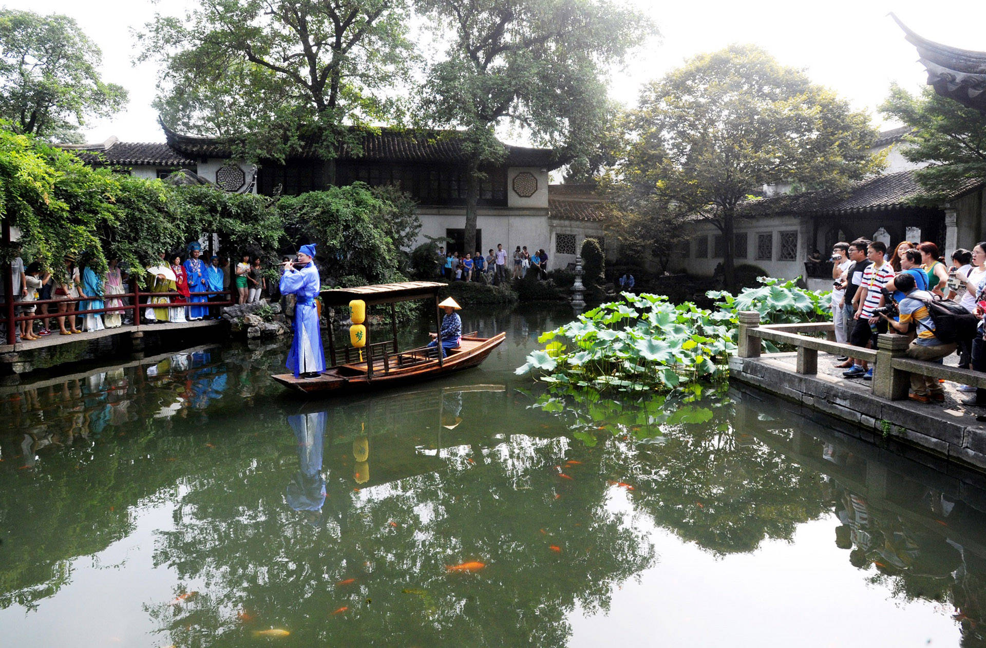 Suzhou 桌布 壁紙 — 一個傳統中式花園池塘，有一艘木船載著一位穿藍袍的人和一位戴錐形帽的人，周圍有石橋上的遊客和茂密的綠植。