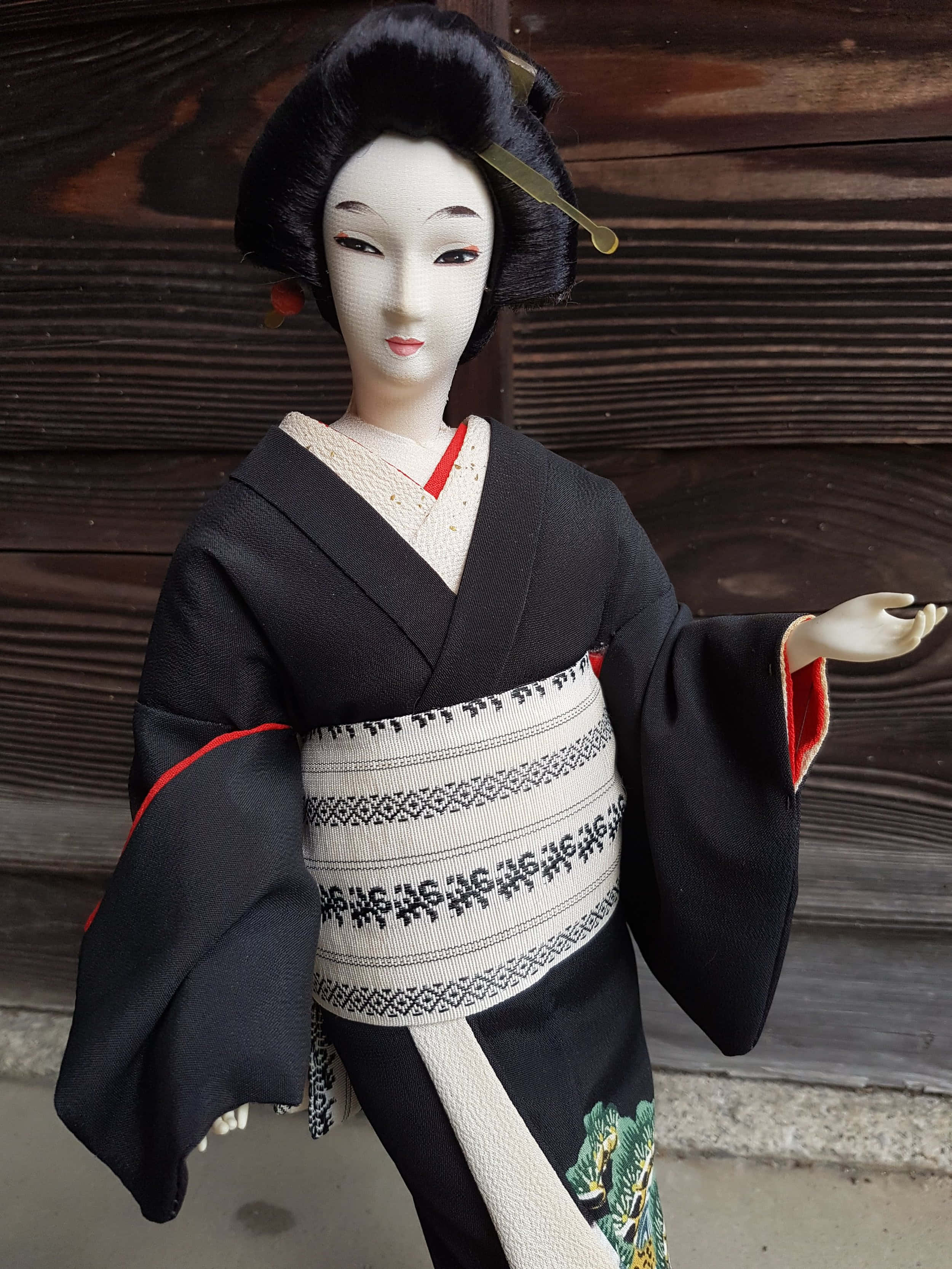 Doll 桌布 壁紙 — 一個傳統日本藝妓娃娃，身穿黑色和服，配有紅色點綴和圖案腰帶，站在深色木牆前。