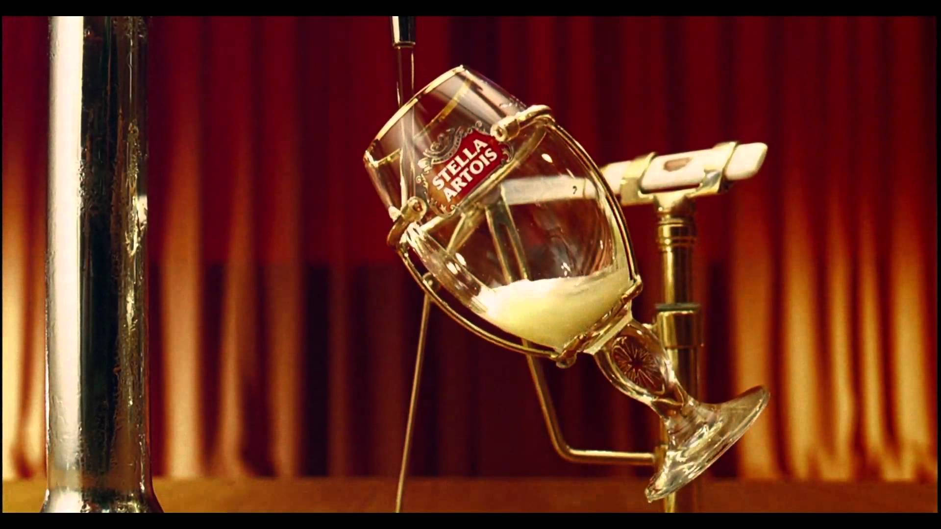Drink 桌布 壁紙 — 一個傾斜在龍頭上的 Stella Artois 啤酒杯，裝滿了金色啤酒和白色泡沫，背景是濃郁的紅色窗簾。
