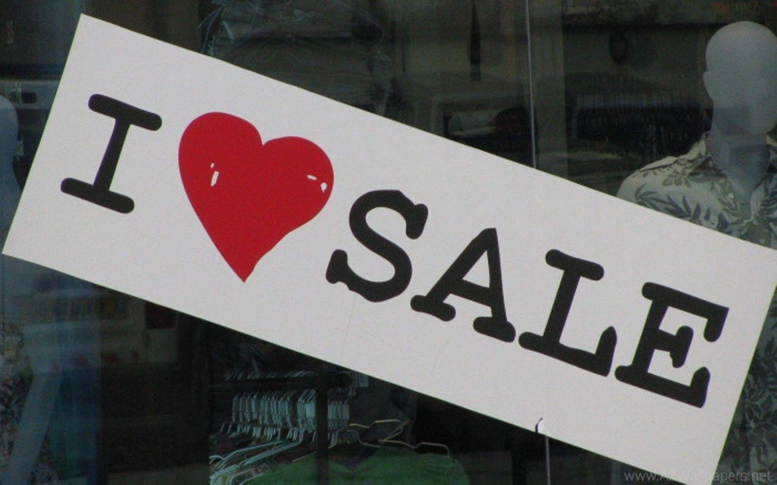 Sales 桌布 壁紙 — 一個傾斜的白色標牌，上面有粗體黑色文字寫著「I ❤️ SALE」，並有一個紅色心形，展示在商店櫥窗上，後面可見人體模型和衣架。