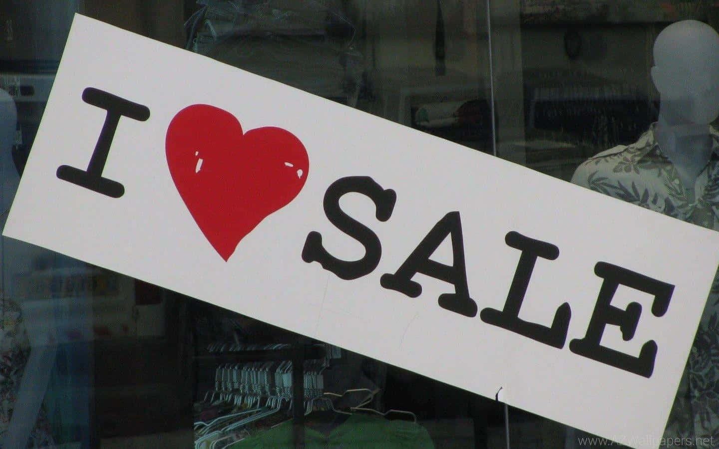 Sales 桌布 壁紙 — 一個傾斜的白色標牌，上面有粗體黑色文字寫著「I ❤️ SALE」，帶有一個紅色心形，展示在商店櫥窗中，後面可見人體模型和衣架。