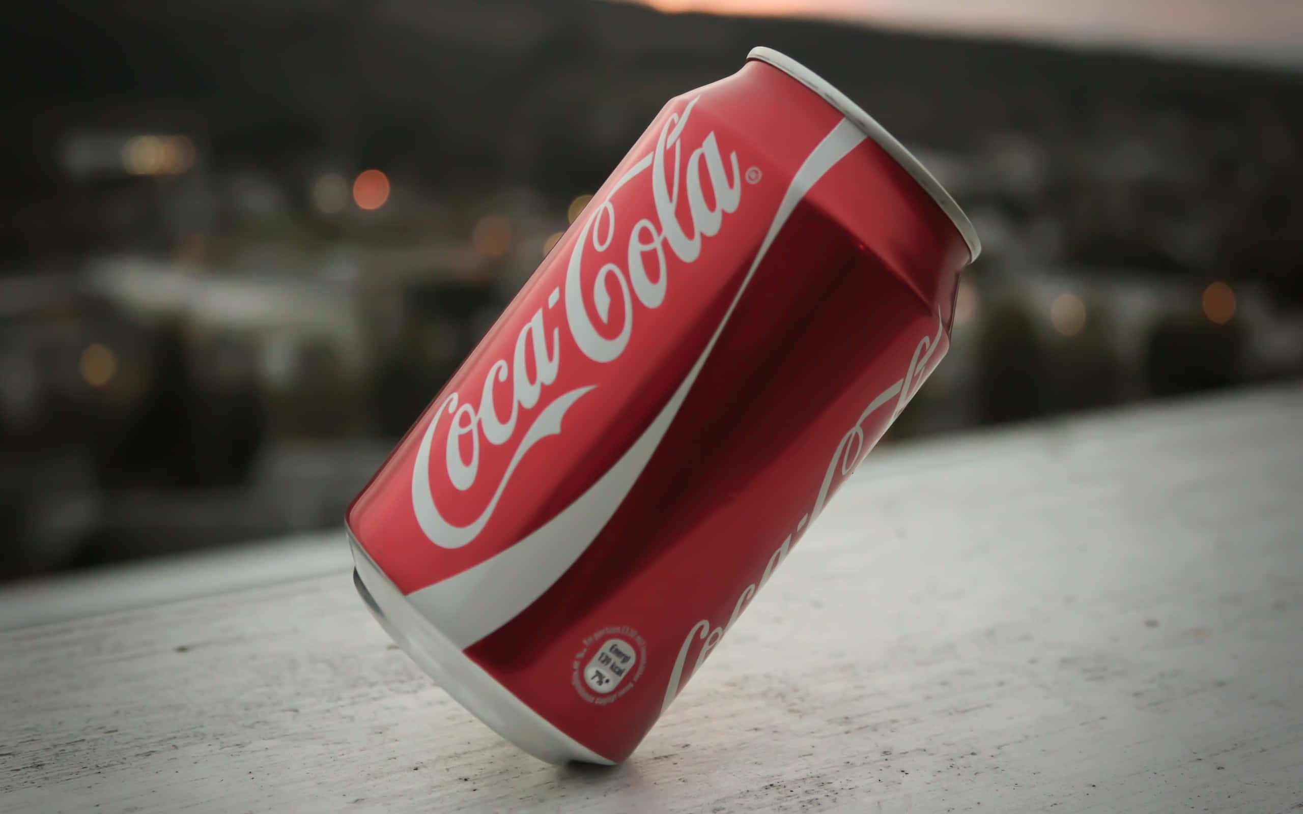 coca cola 桌布 壁紙 — 一個傾斜的紅色可口可樂罐放在一個風化的白色表面上，背景是黃昏時模糊的城市景觀。