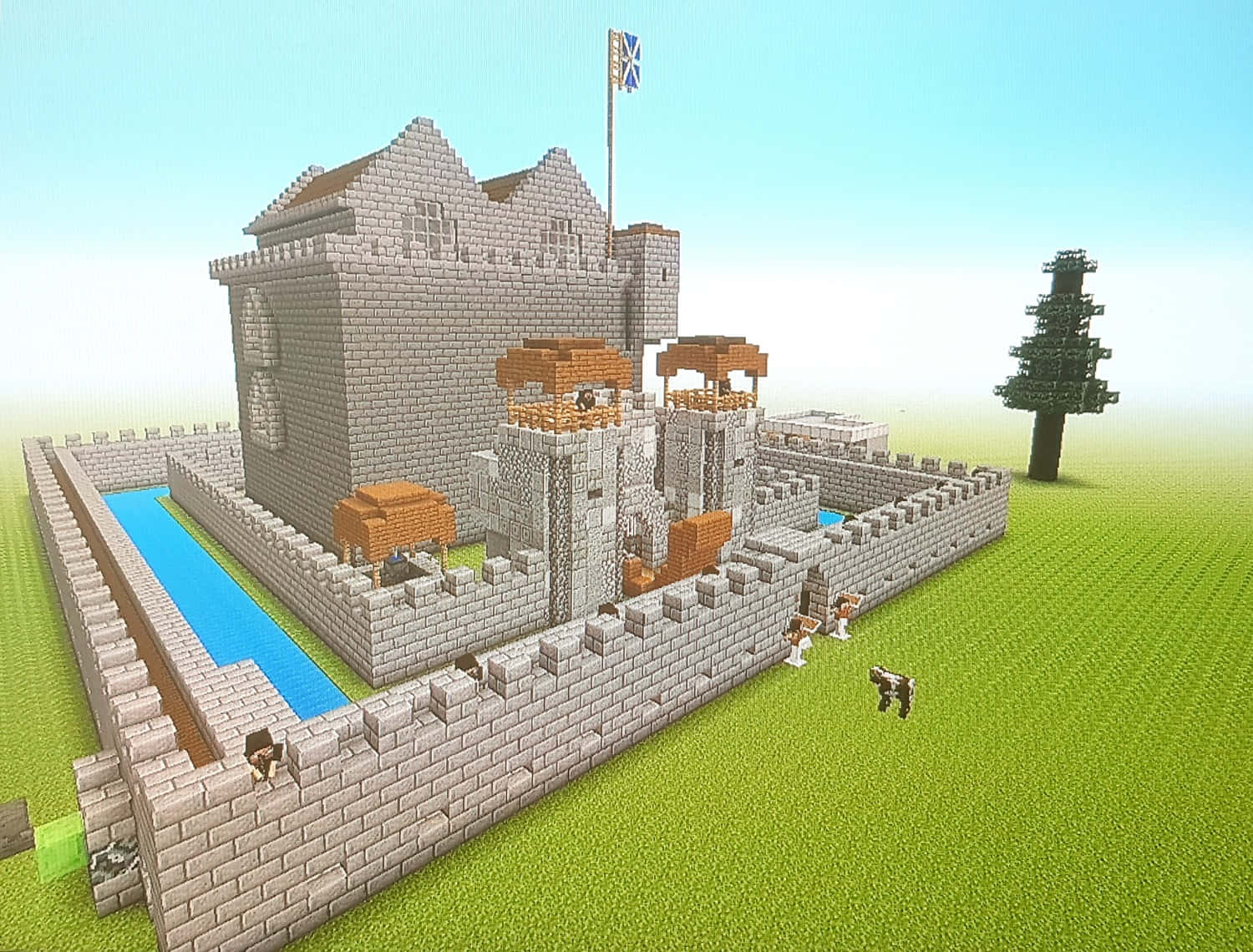 Minecraft Castles 桌布 壁紙 — 一個像素化的Minecraft城堡，有石牆、護城河和旗幟，矗立在綠色草地上，旁邊有一棵孤樹。