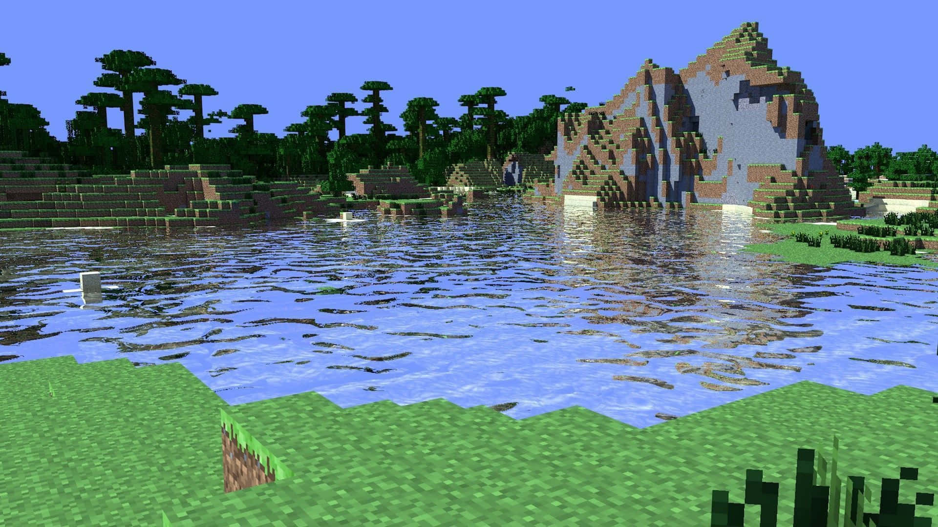 Minecraft Survival 桌布 壁紙 — 一個像素化的Minecraft風景，平靜的湖面倒映著岩石懸崖和森林，天空清澈湛藍。