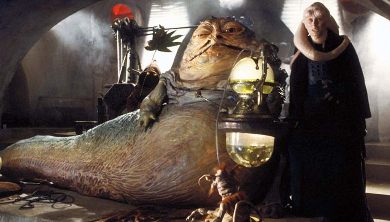 A New Hope 桌布 壁紙 — 一個像蛞蝓的大型外星人躺在一間燈光昏暗、雜亂的房間裡，而一個長頸的高大人形生物站在他旁邊，手持一盞發光的燈。