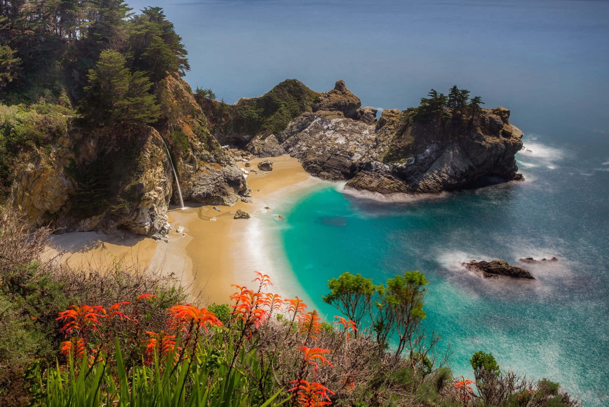 Big Sur 桌布 壁紙 — 一個僻靜的海灣，碧綠的海水環繞著金色沙灘，沙灘兩旁是崎嶇的懸崖和鮮豔的橙色花朵。