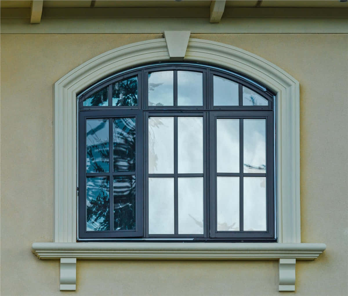 window 桌布 壁紙 — 一個優雅的拱形窗戶，帶有深色框架和白色飾邊，設置在米色灰泥牆上，反射出多雲的天空和樹枝。