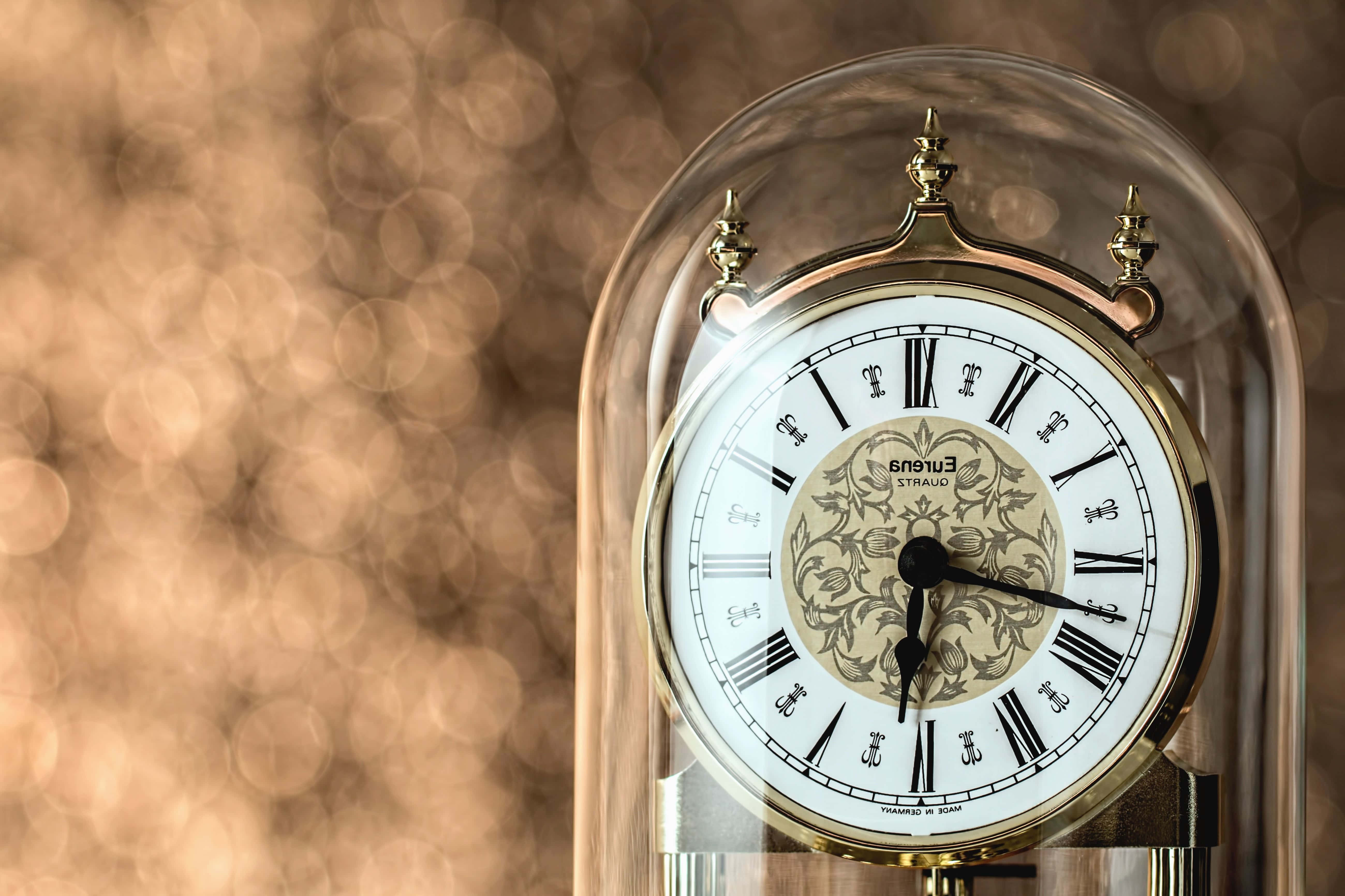 Clock 桌布 壁紙 — 一個優雅的黃銅鐘，帶有羅馬數字和華麗花卉中心，置於玻璃罩下，背景是溫暖的散景效果。