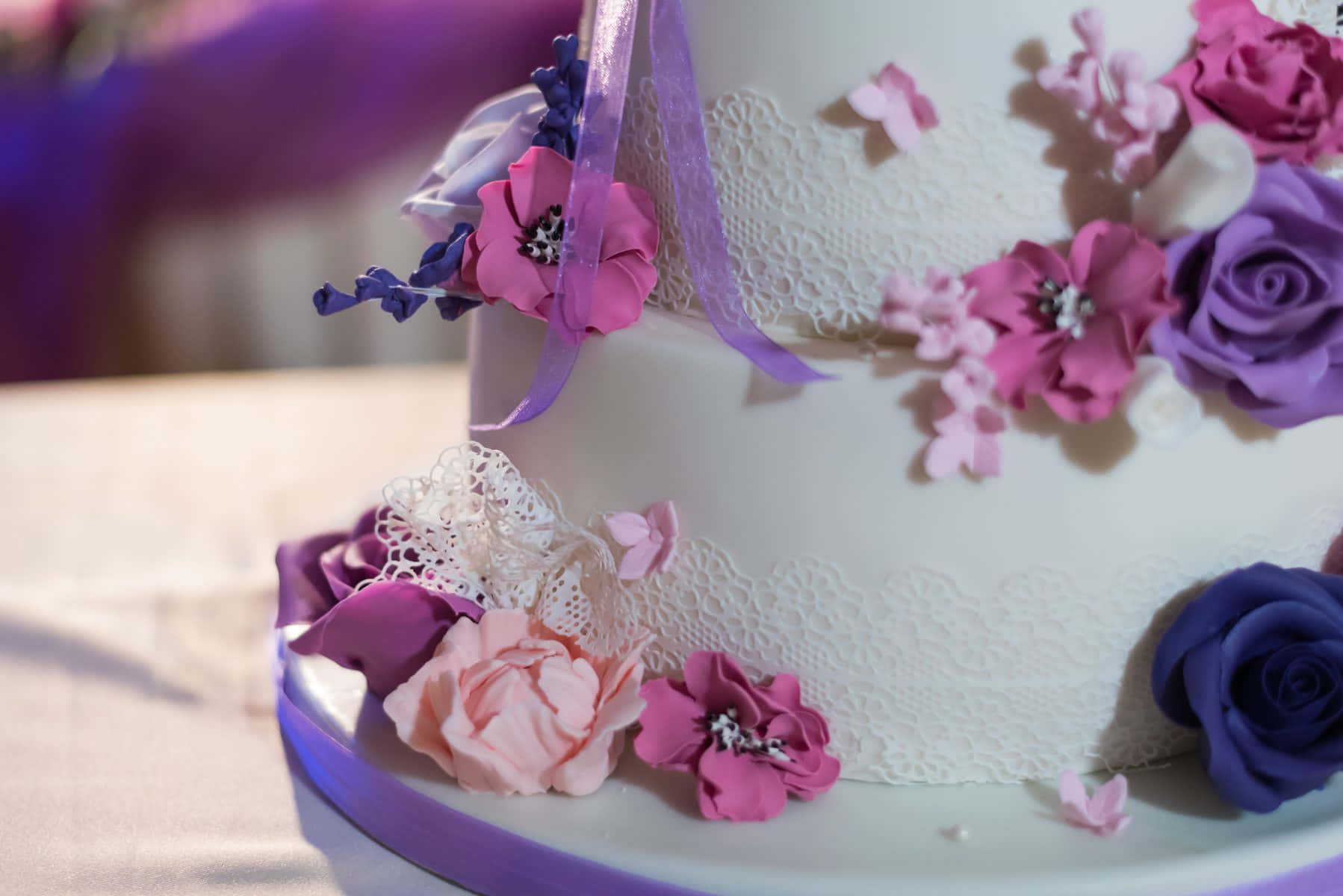 Cake Fondant 桌布 壁紙 — 一個優雅裝飾的婚禮蛋糕近距離特寫，飾有粉紅色、紫色和藍色的糖花、蕾絲和絲帶。
