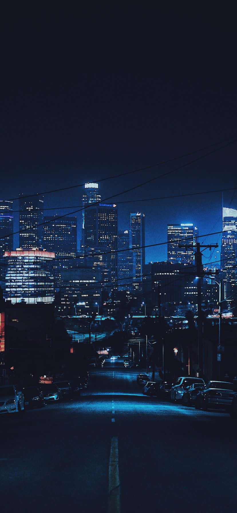 桌布 壁紙 — 一個充滿氛圍的夜景城市景觀，發光的摩天大樓和一條安靜的街道，兩旁停滿了汽車，在深藍色的天空下。