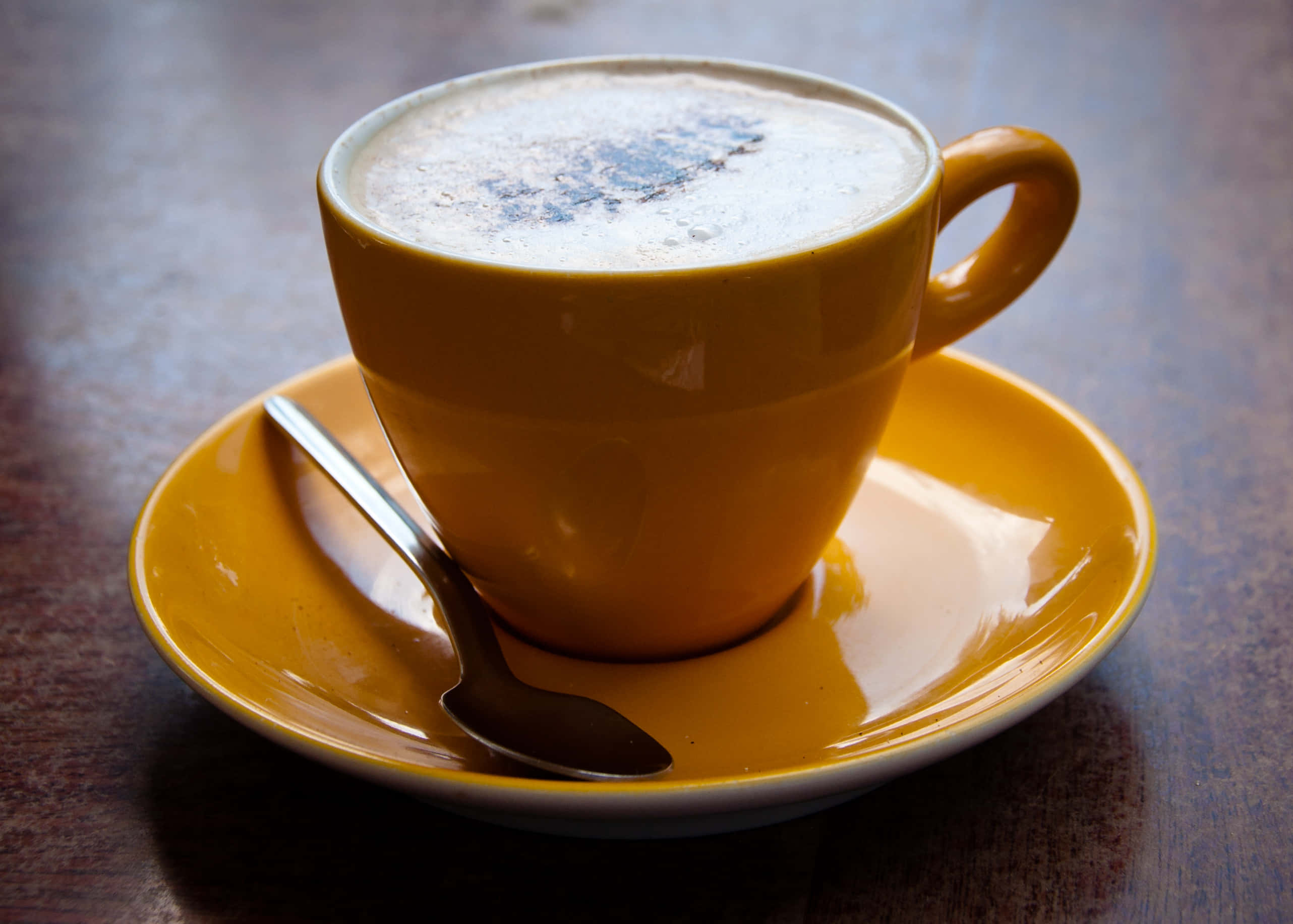 Coffee Cup 桌布 壁紙 — 一個充滿泡沫卡布奇諾的鮮黃色陶瓷杯，放在配套的碟子上，旁邊有一隻湯匙，置於深色木桌上。