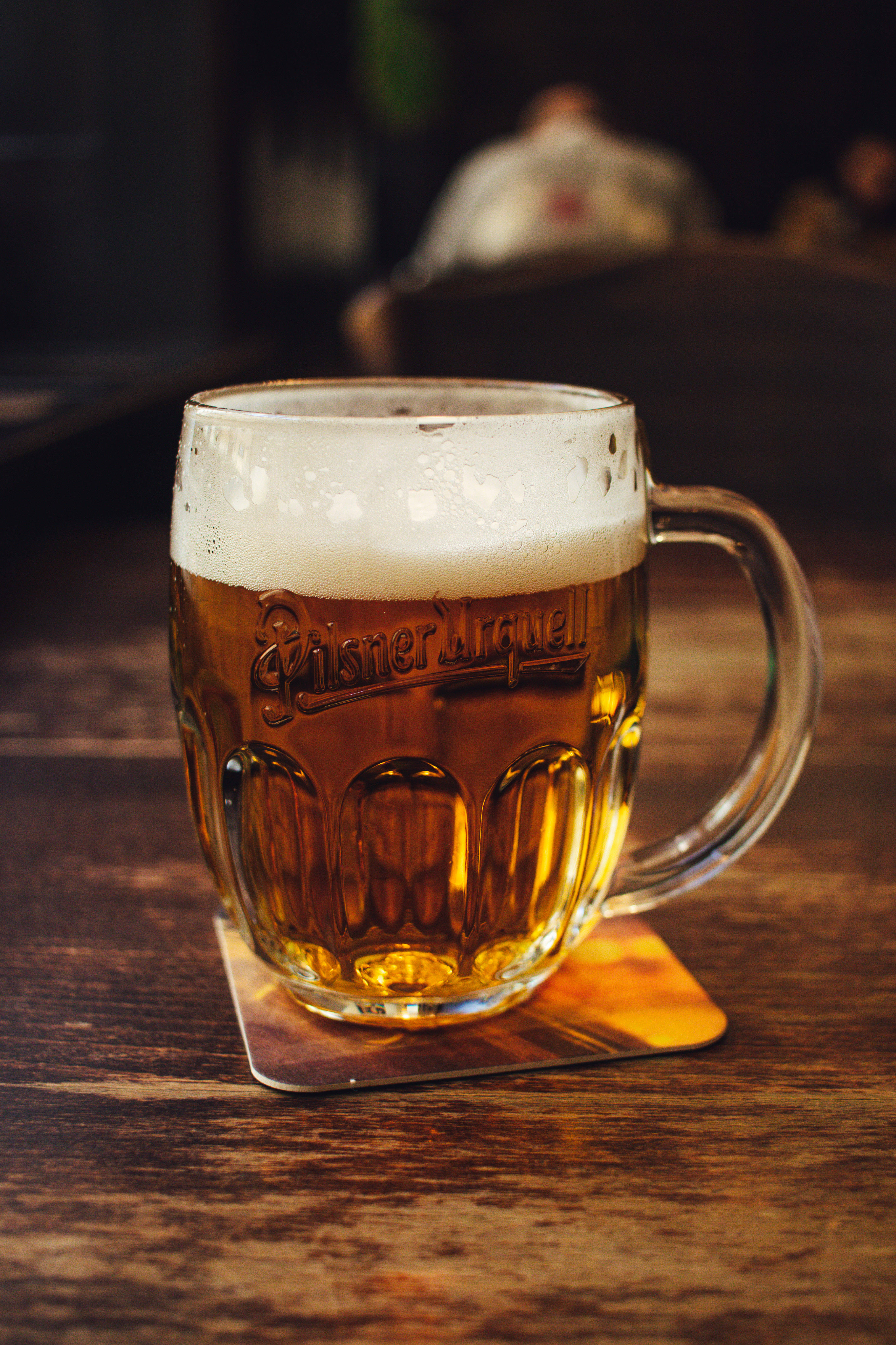 Beer 桌布 壁紙 — 一個充滿泡沫的 Pilsner Urquell 啤酒杯放在昏暗酒吧的木桌上，背景有一個模糊的顧客。