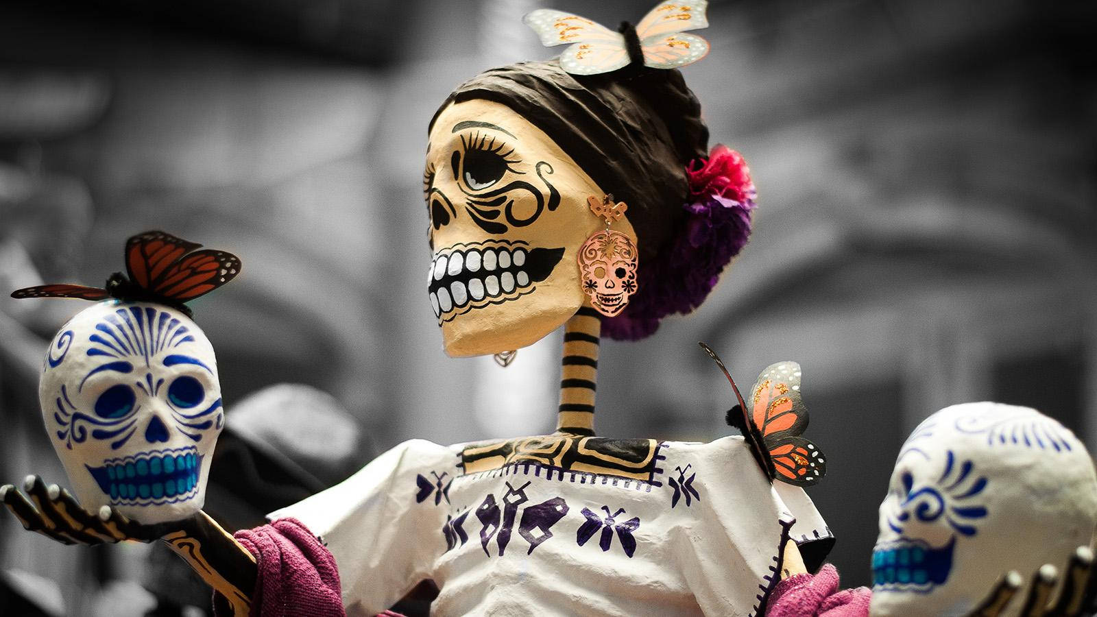 Dia De Los Muertos 桌布 壁紙 — 一個充滿活力的 papel picado 骷髏人物，飾有蝴蝶和花卉頭飾，在模糊的城市背景下手持裝飾過的頭骨。