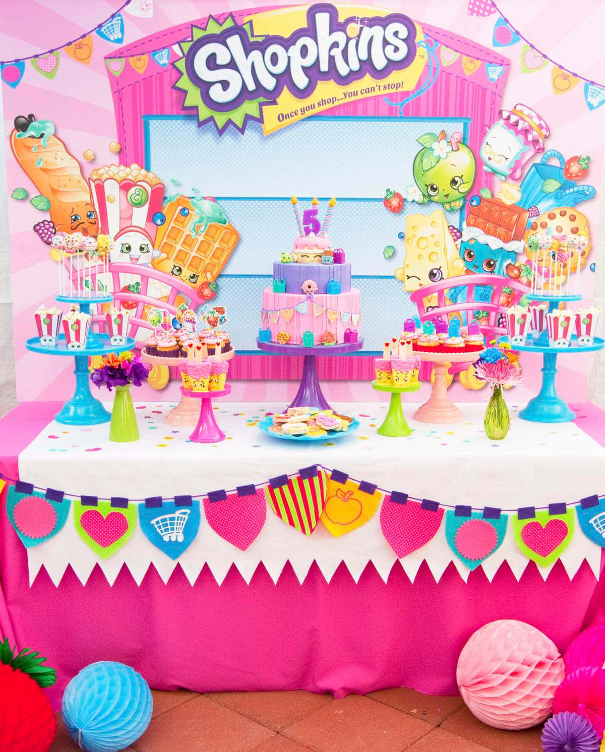 Shopkins 桌布 壁紙 — 一個充滿活力的Shopkins主題生日派對桌，擺放著色彩繽紛的蛋糕、角色裝飾和節日彩旗，在明亮的日光下。