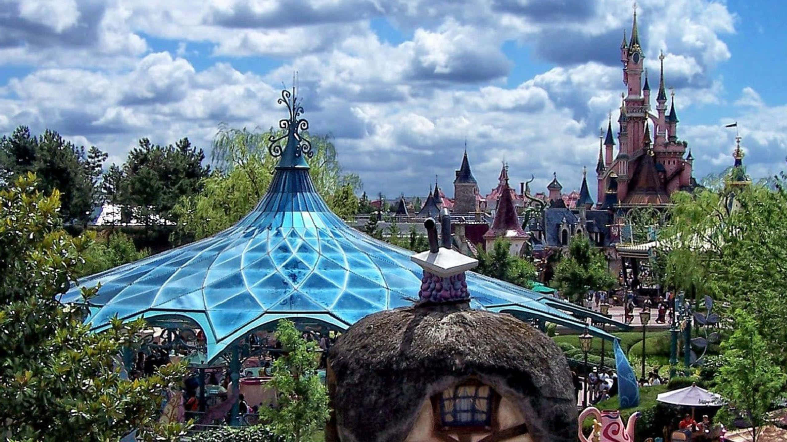Disneyland 桌布 壁紙 — 一個充滿活力的主題公園場景，前景是藍色玻璃圓頂結構，背景是粉紅色城堡，天空多雲。