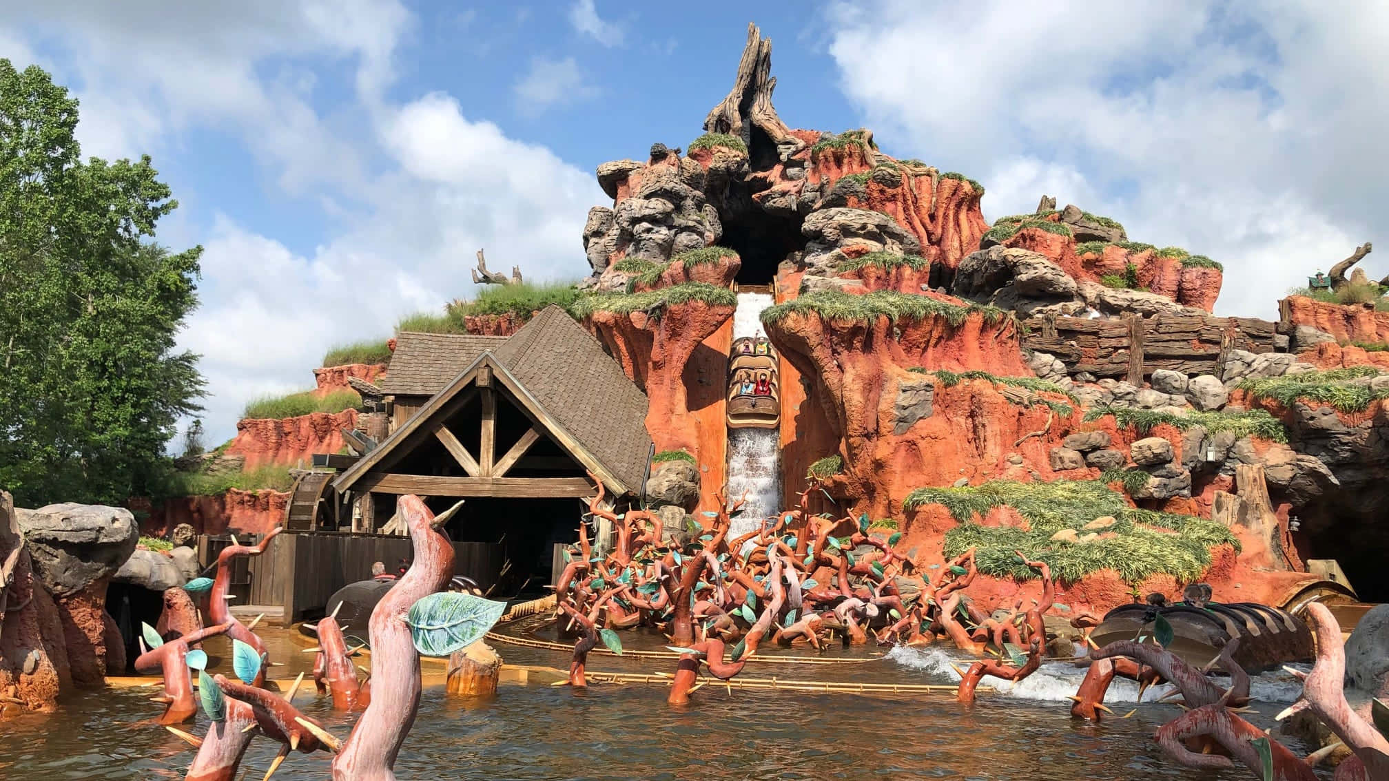 Splash Mountain 桌布 壁紙 — 一個充滿活力的主題公園景點，木製建築坐落在紅色岩石懸崖中，水中擺放著奇特的樹狀雕塑，在明亮的藍天下。