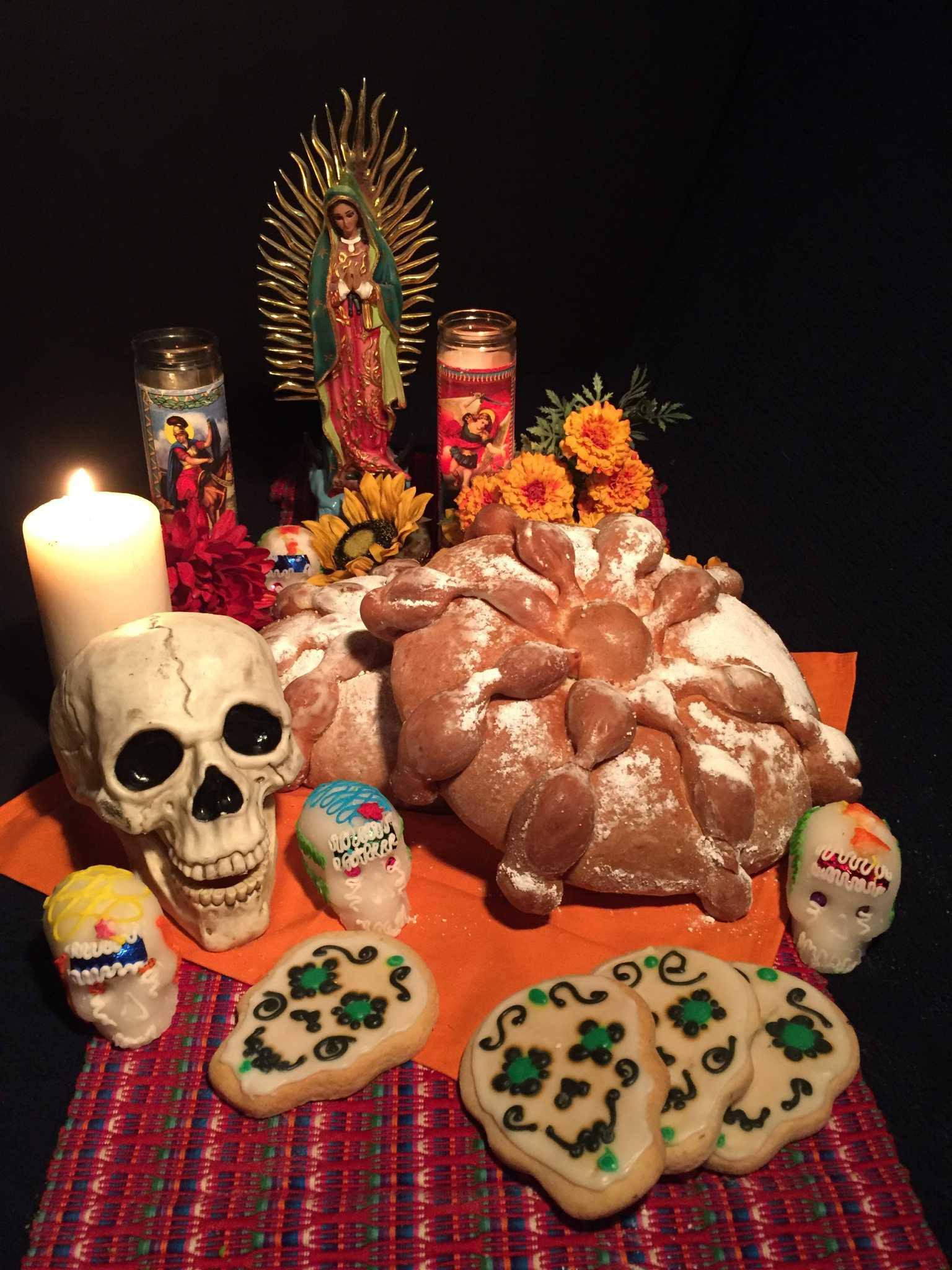 Mexican 桌布 壁紙 — 一個充滿活力的亡靈節祭壇，上面有糖骷髏、麵包、蠟燭和金盞花，排列在黑色背景前的彩色布上。