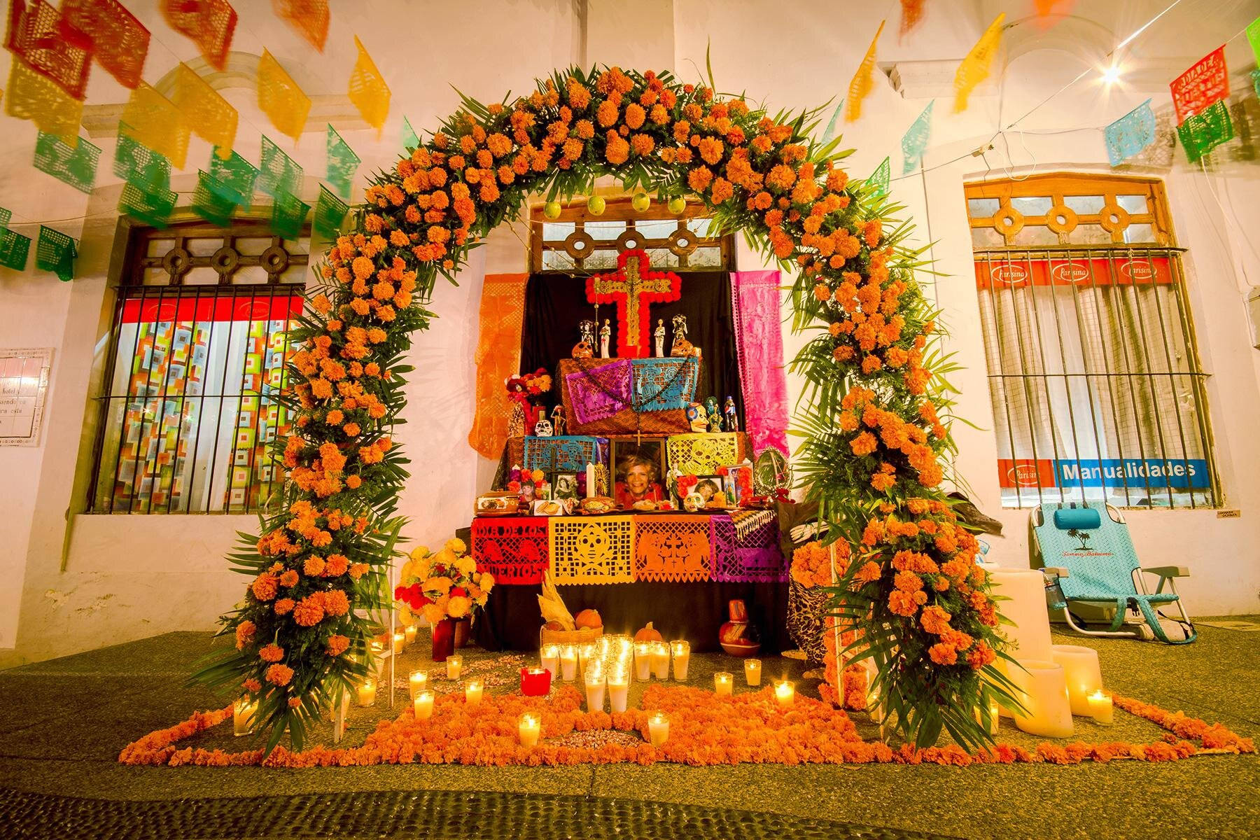 Mexican 桌布 壁紙 — 一個充滿活力的亡靈節祭壇，裝飾著橙色萬壽菊、蠟燭和色彩繽紛的 papel picado，在溫暖的室內燈光下。