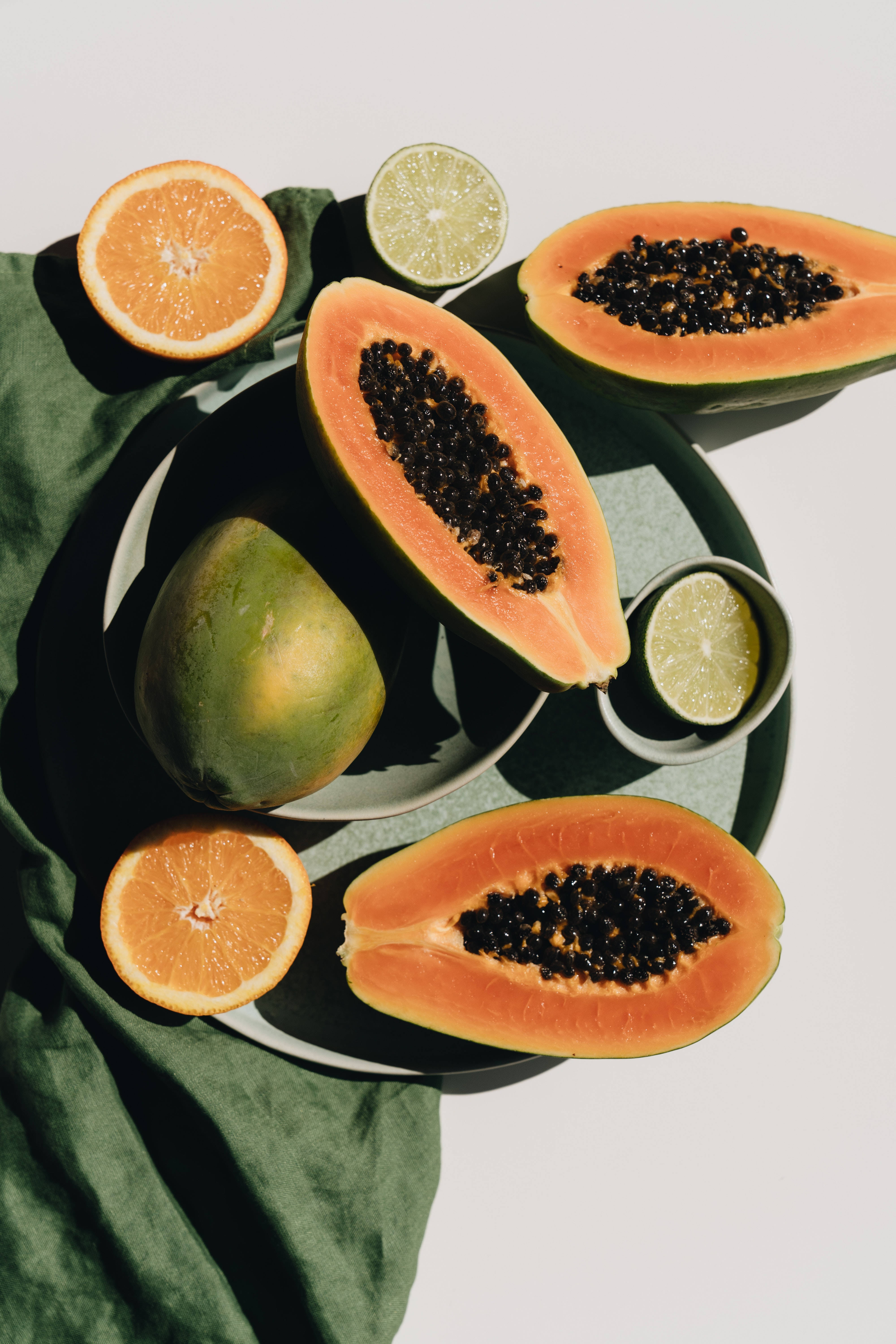 Fruits 桌布 壁紙 — 一個充滿活力的俯拍鏡頭，展示切片木瓜、柳橙和萊姆放在綠色布料上，帶有強烈陰影。