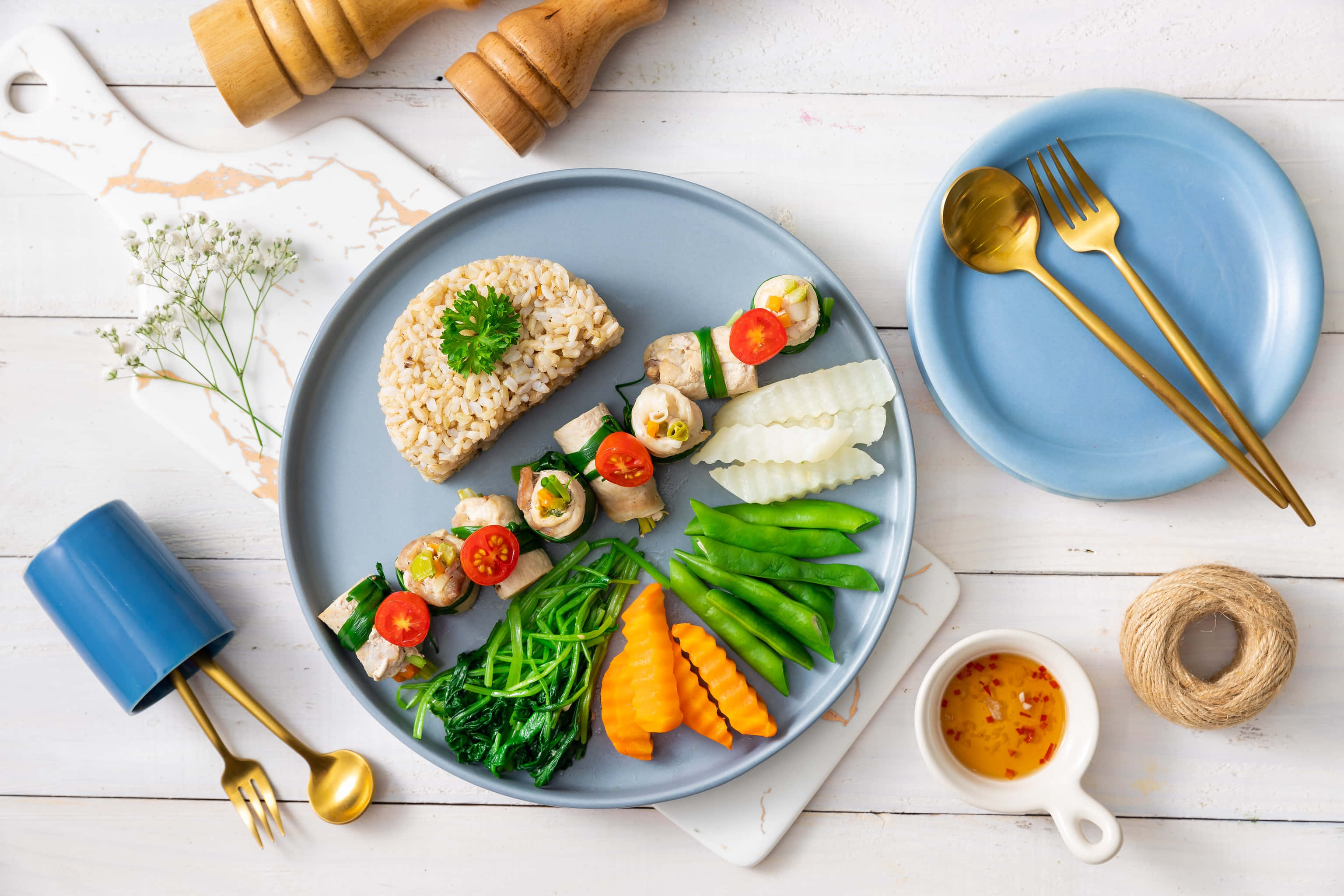 Healthy Food 桌布 壁紙 — 一個充滿活力的健康餐，有米飯、雞肉卷、蔬菜和醬汁，藝術地擺放在藍色盤子上，配金色餐具，放在白色木桌上。