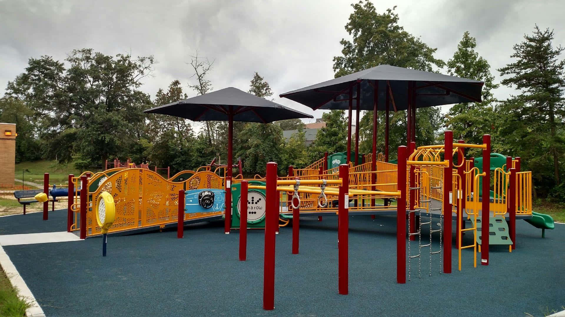 Playground 桌布 壁紙 — 一個充滿活力的兒童遊樂場，設有黃色和紅色的遊樂設施，黑色遮陽傘下，藍色安全地面上，周圍樹木環繞。
