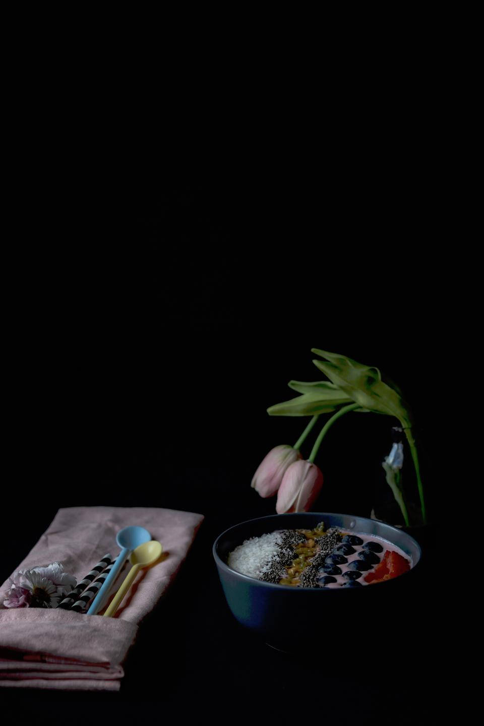 Fruits 桌布 壁紙 — 一個充滿活力的冰沙碗，上面放有奇亞籽和水果，旁邊有粉紅色的鬱金香和色彩繽紛的湯匙，背景是純黑色。