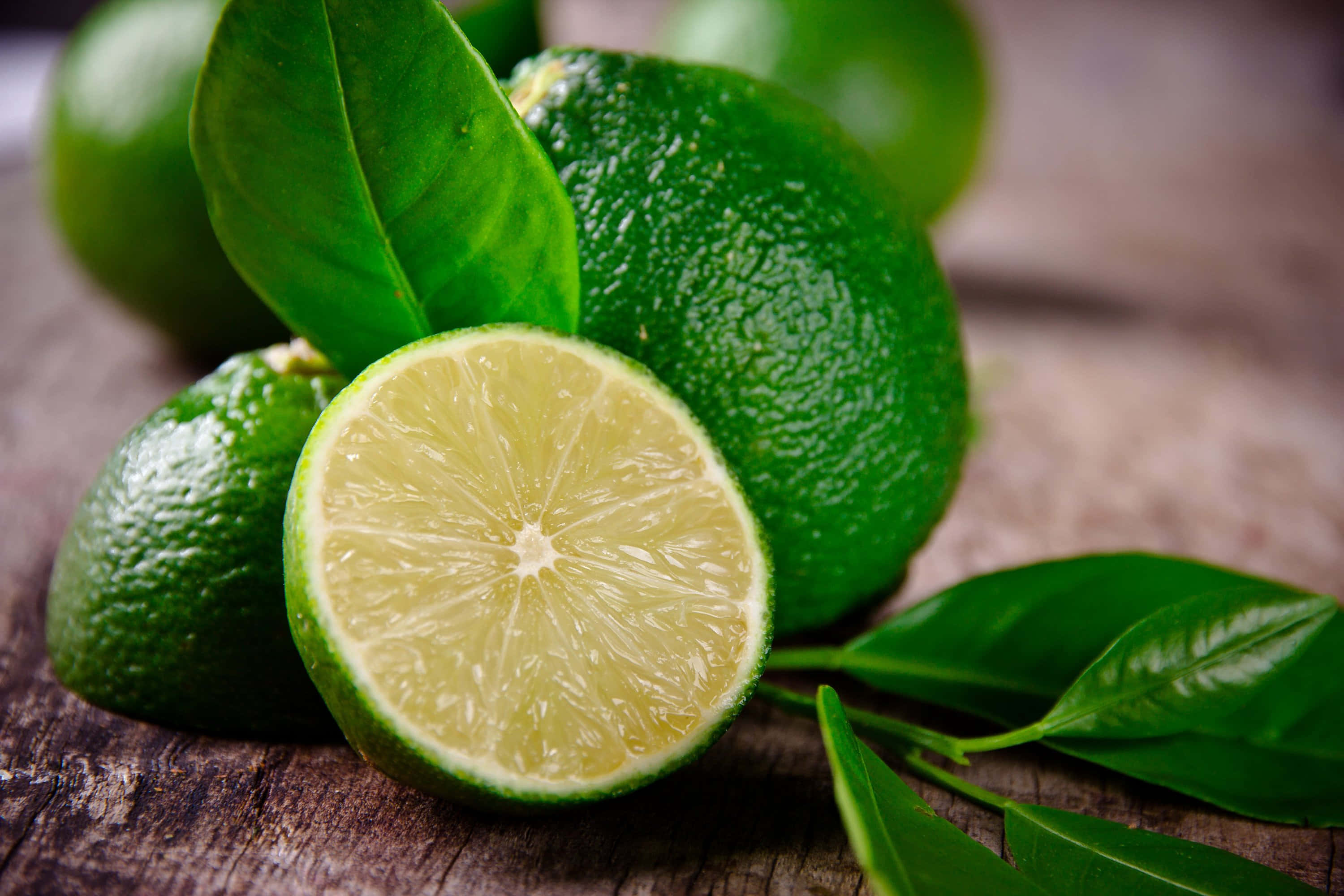Lime 桌布 壁紙 — 一個充滿活力的半切萊姆，帶有多汁的果肉，與整個萊姆和光澤的葉子一起放在質樸的木質表面上。
