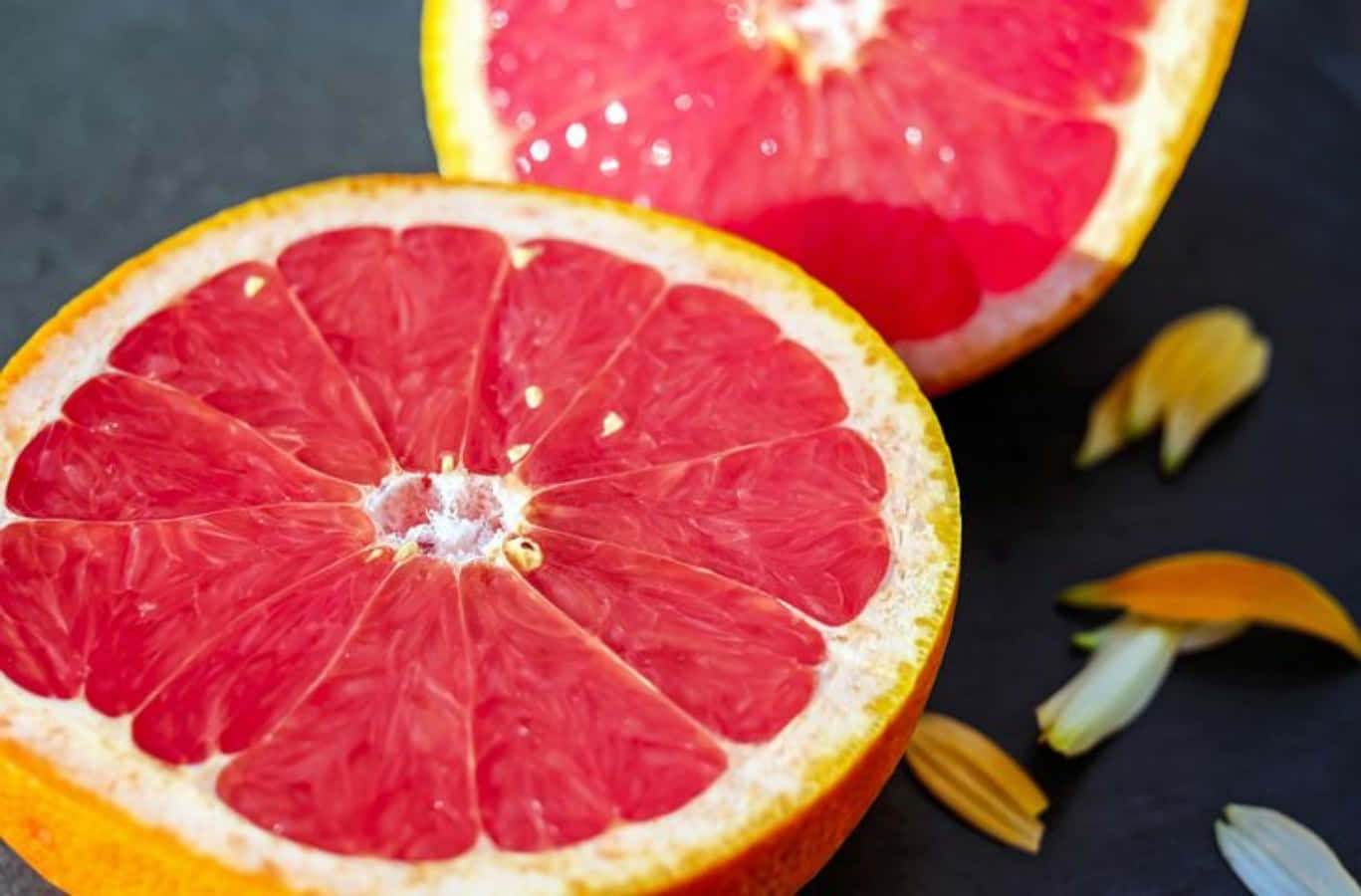 Pink Grapefruit 桌布 壁紙 — 一個充滿活力的半切葡萄柚在深色石板表面上露出多汁的粉紅色果肉，周圍散落著花瓣。