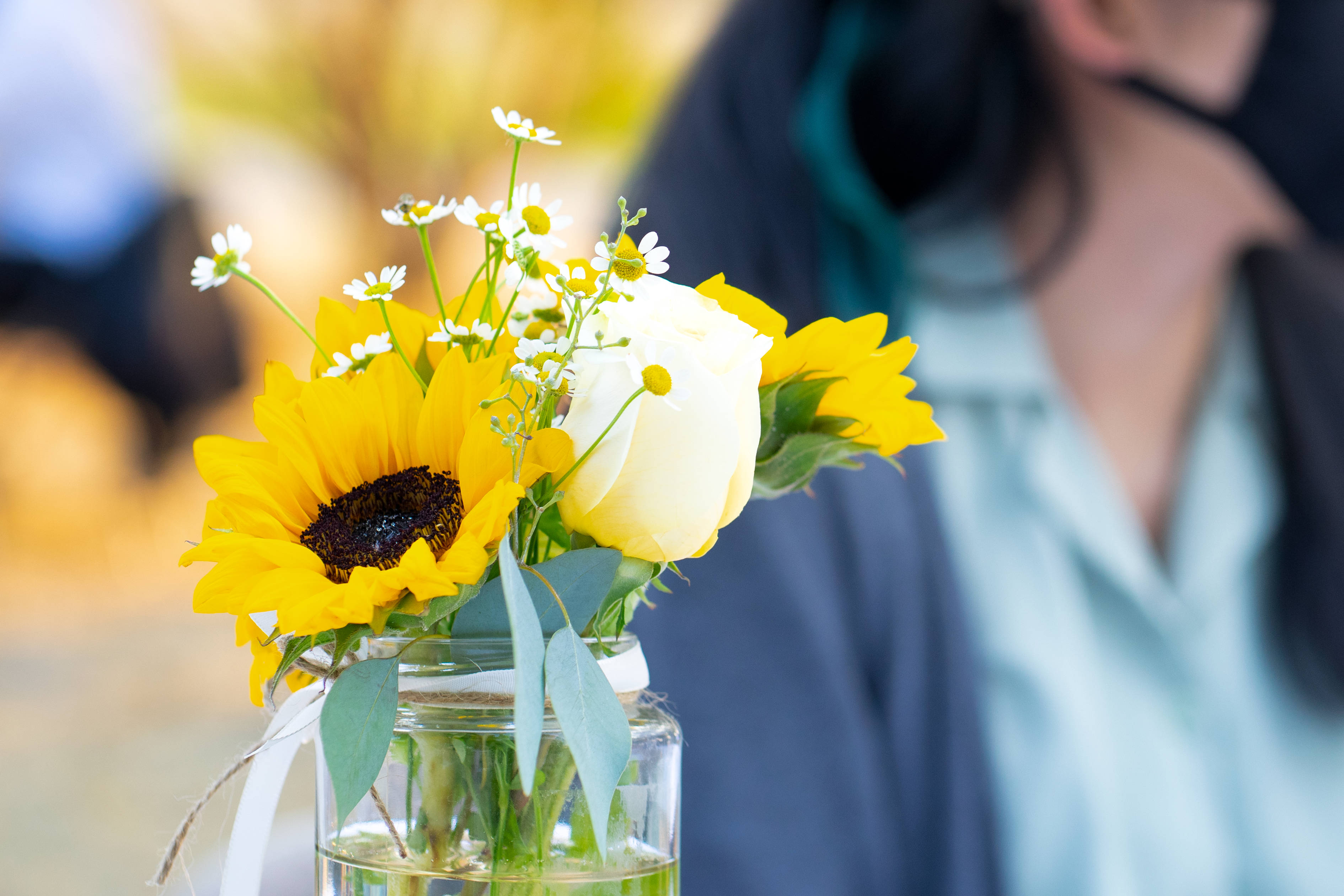 Sunflower Aesthetic 桌布 壁紙 — 一個充滿活力的向日葵和白玫瑰花束，放在玻璃罐中，搭配尤加利葉，柔和對焦在一個穿著淺藍色襯衫的模糊人物前。