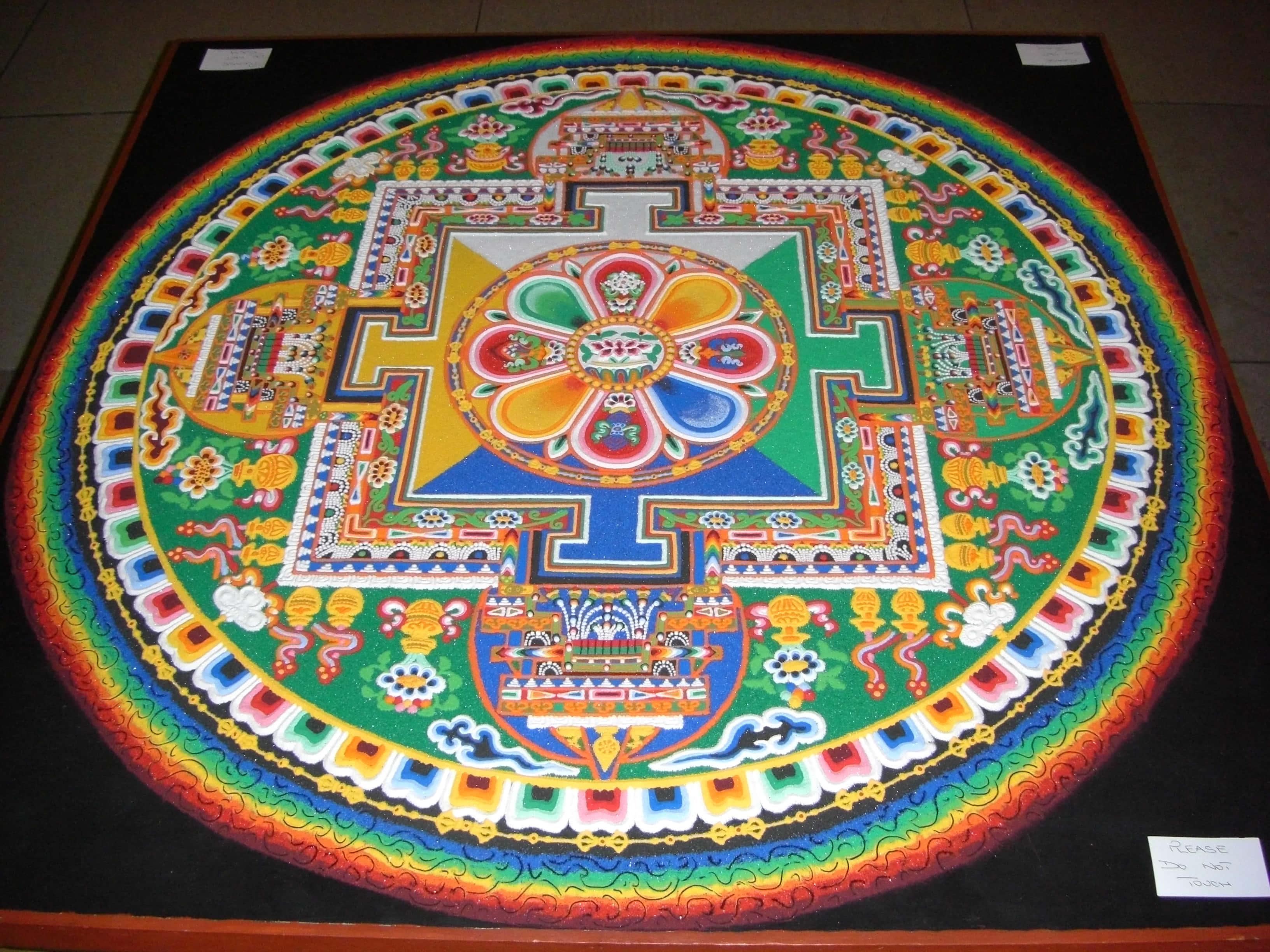 Sand Art 桌布 壁紙 — 一個充滿活力的圓形西藏沙壇城，帶有複雜的幾何圖案和彩虹邊框，放置在深色地板上。