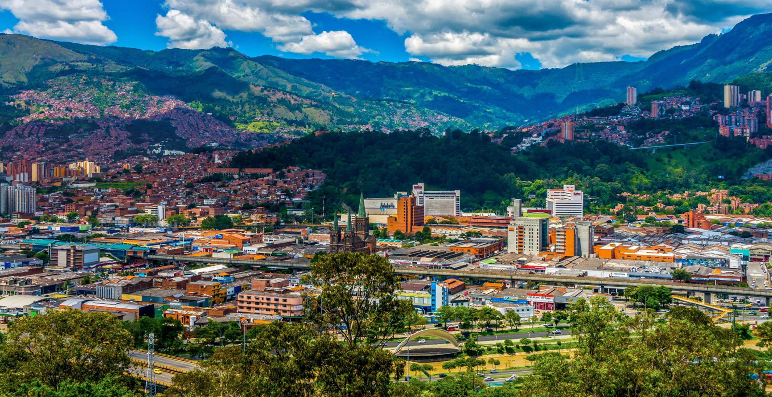 Medellin 桌布 壁紙 — 一個充滿活力的城市，在明亮的藍天和蓬鬆的白雲下，蔓延在鬱鬱蔥蔥的綠色山丘上。