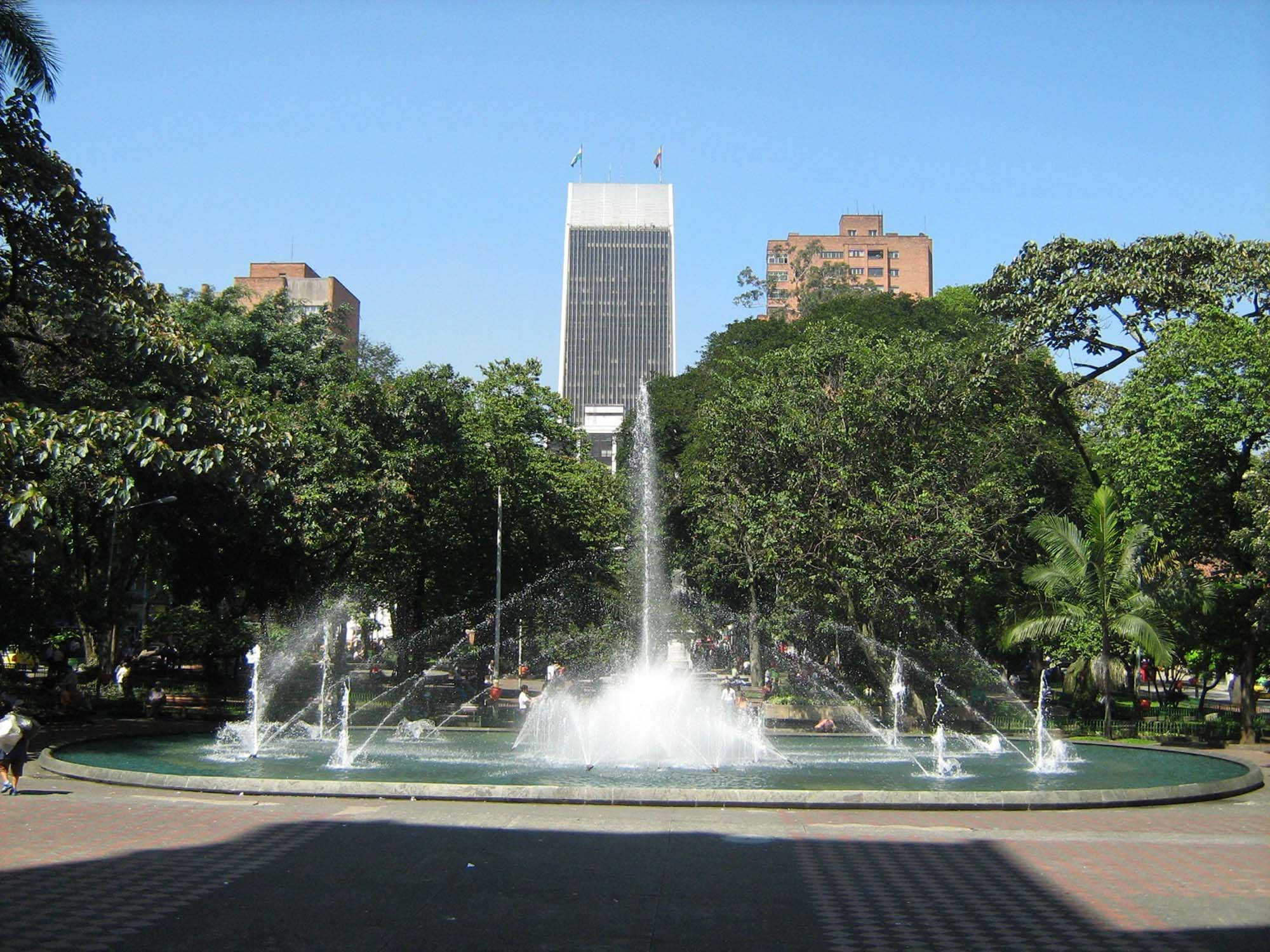 Medellin 桌布 壁紙 — 一個充滿活力的城市公園噴泉在清澈的藍天下噴水，高樓大廈在茂密的綠樹後方聳立。
