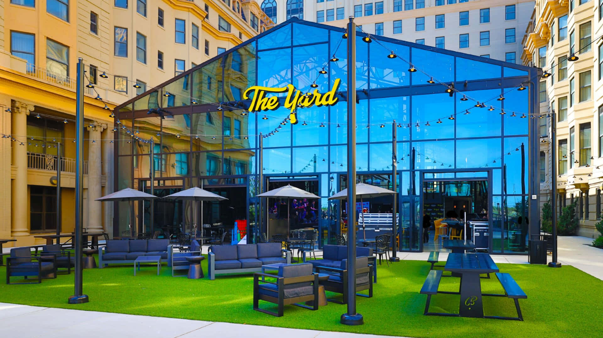 Hotel 桌布 壁紙 — 一個充滿活力的城市庭院，設有玻璃牆的場地「The Yard」，設有綠色草皮野餐桌和燈串下的休息座位。